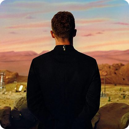 Justin Timberlake in a black suit standing outdoors, facing a desert landscape at sunset.