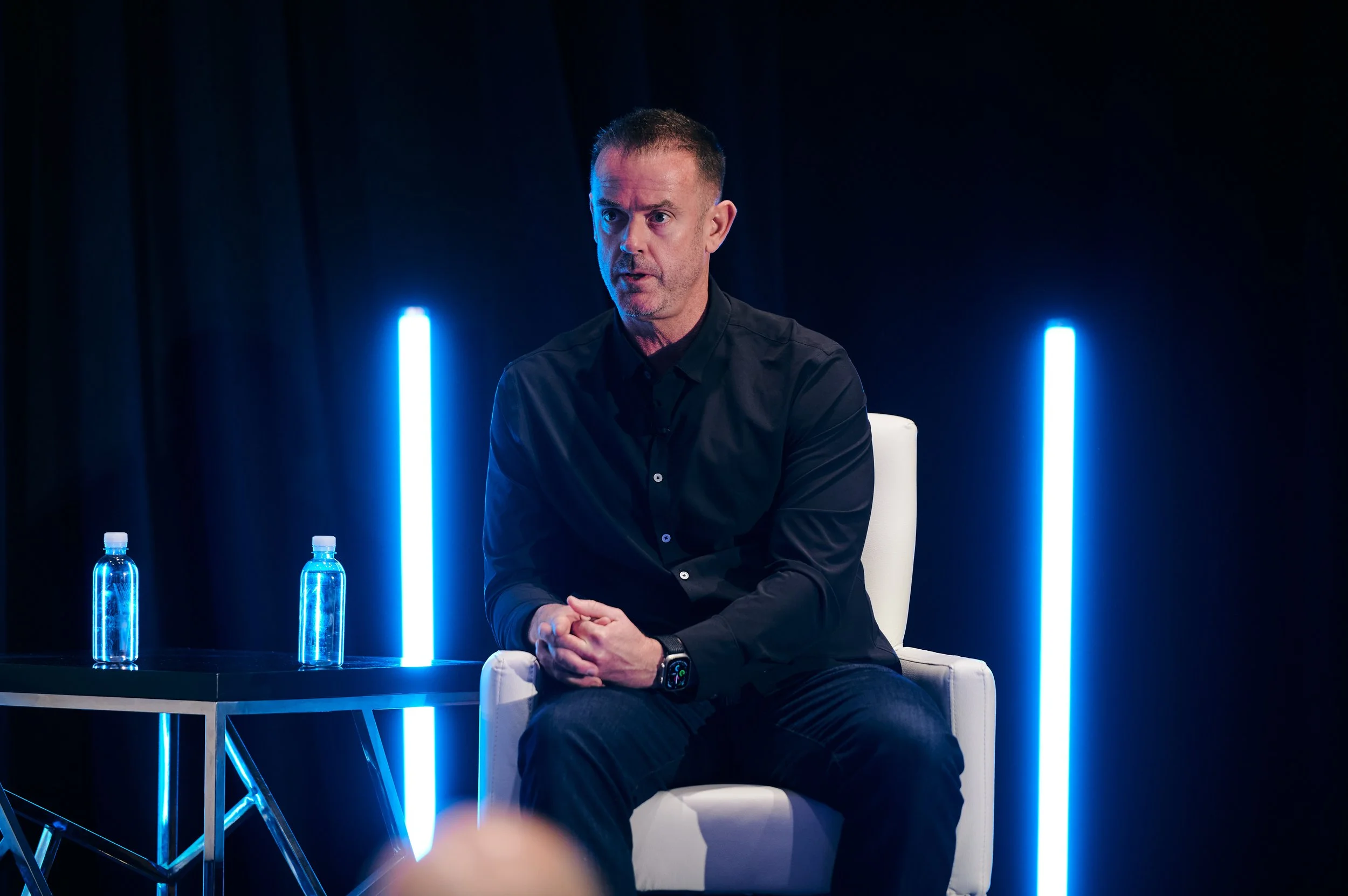 Dustin Engel in a black shirt sitting on a white chair during a panel or interview, with dark background and blue vertical light accents, water bottles on a table beside him.
