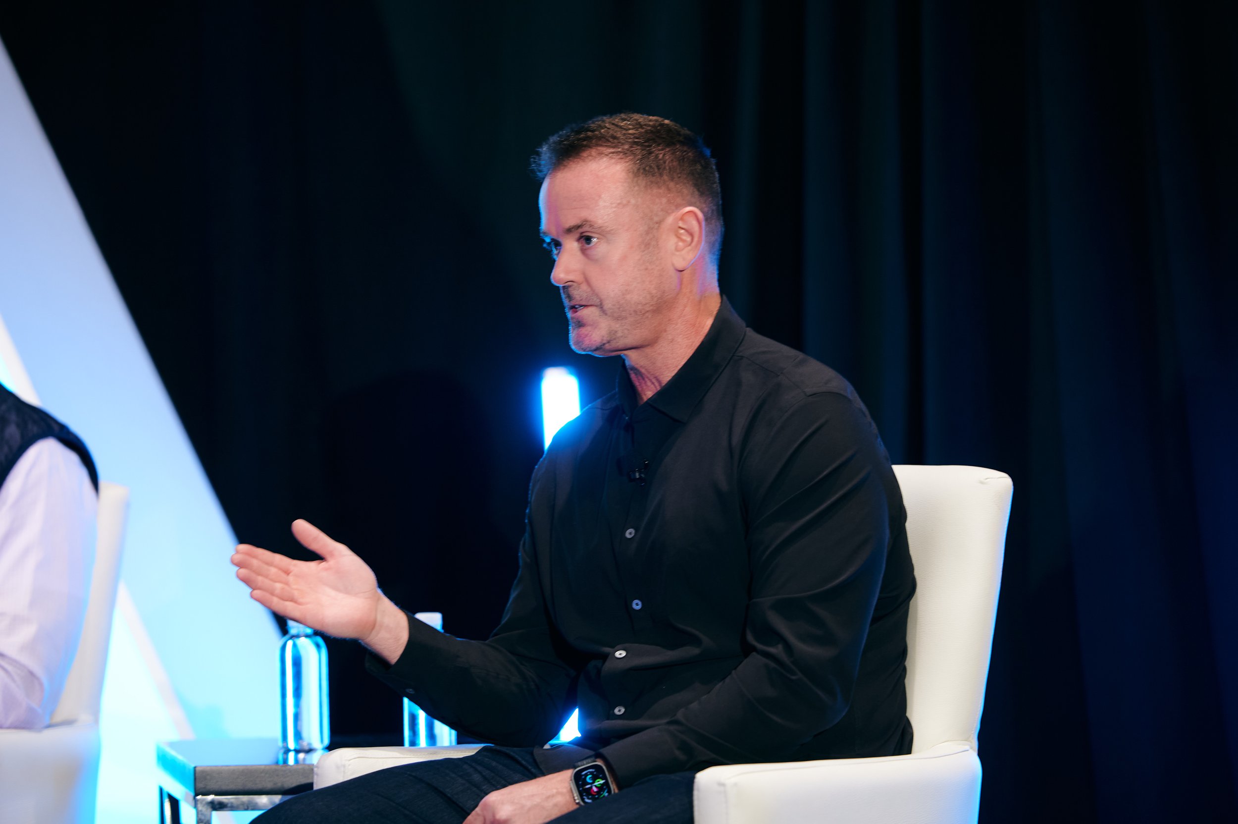 Dustin Engel in a black shirt sitting on a white chair, engaged in conversation, with a dark background and blue lighting.