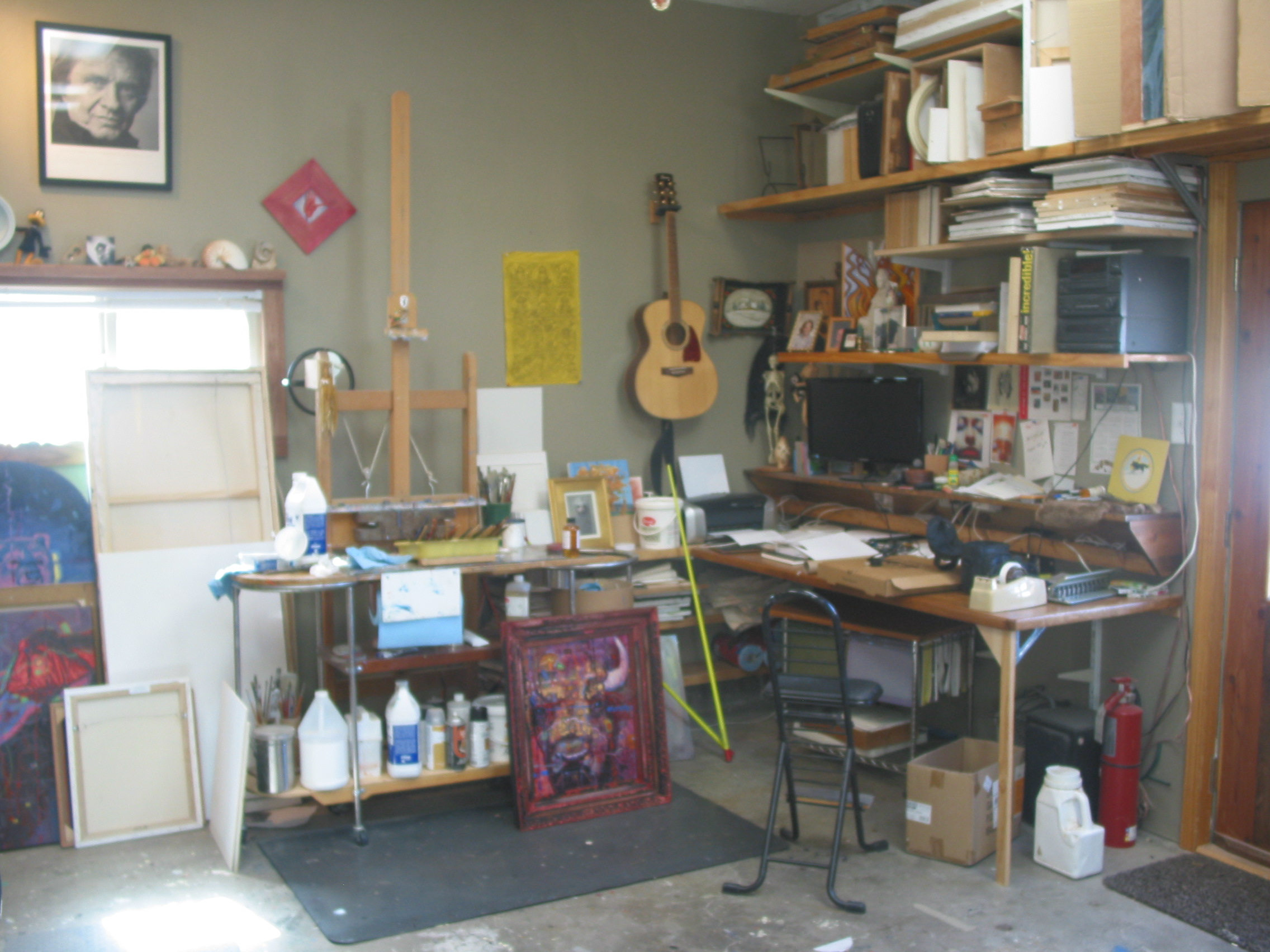 Here is a view of my easel without a painting on it. My Painting Table that holds my pallet and on the right a table and a computer screen that I view my photos and references with. You can see the guitar on the wall and a poster of the Man in Black. There are unfinished canvases on the floor to the left.