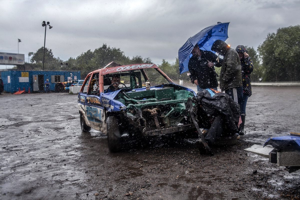 Jack Coveney sits inside his car, whilst undergoes repairs between race heats. Jack is currently in second place in the 2014 PRI Junior Banger championship, His dad aggressively works away at tightening the bolts on the front wheel of the battered car.