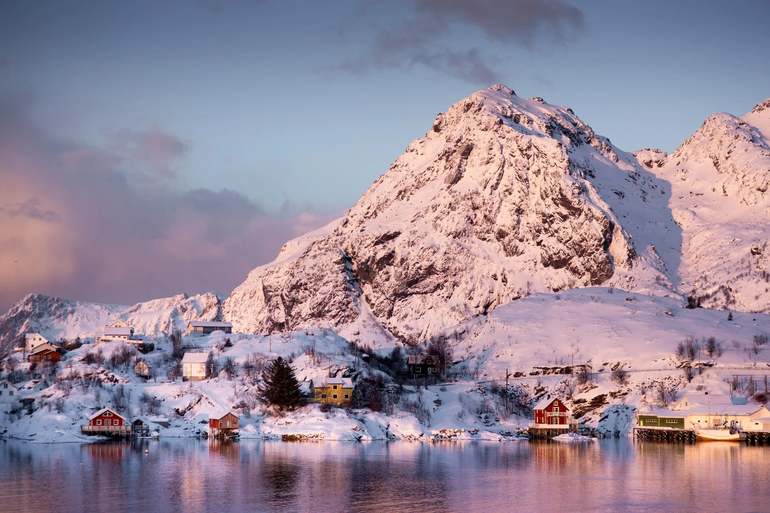 Snow-covered houses along a shoreline with a large snow-covered mountain in the background under a partly cloudy sky.