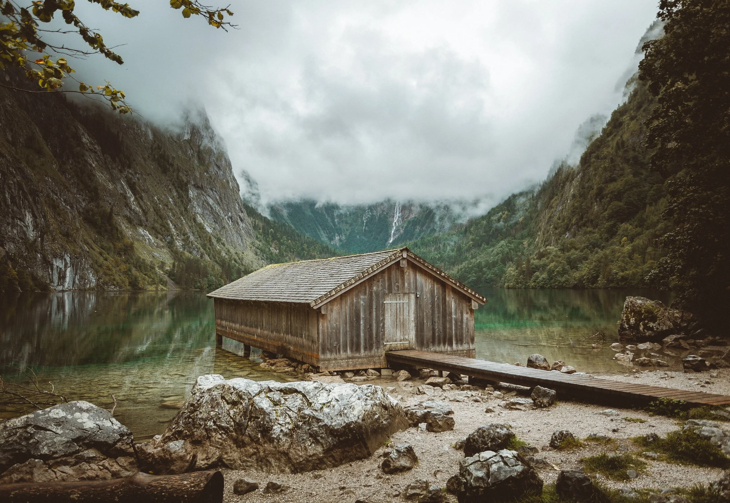 A rustic wooden boathouse on the edge of a calm lake surrounded by green mountains, with cloudy skies and mist.