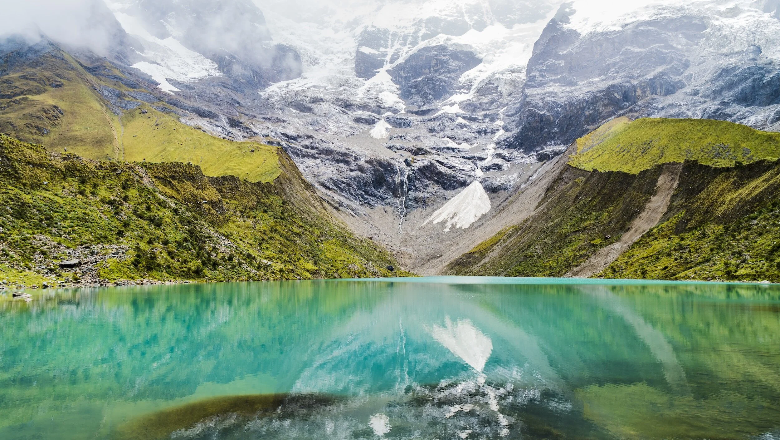 A mountain landscape with a turquoise lake in the foreground, green slopes on either side, and snow-capped peaks in the background.