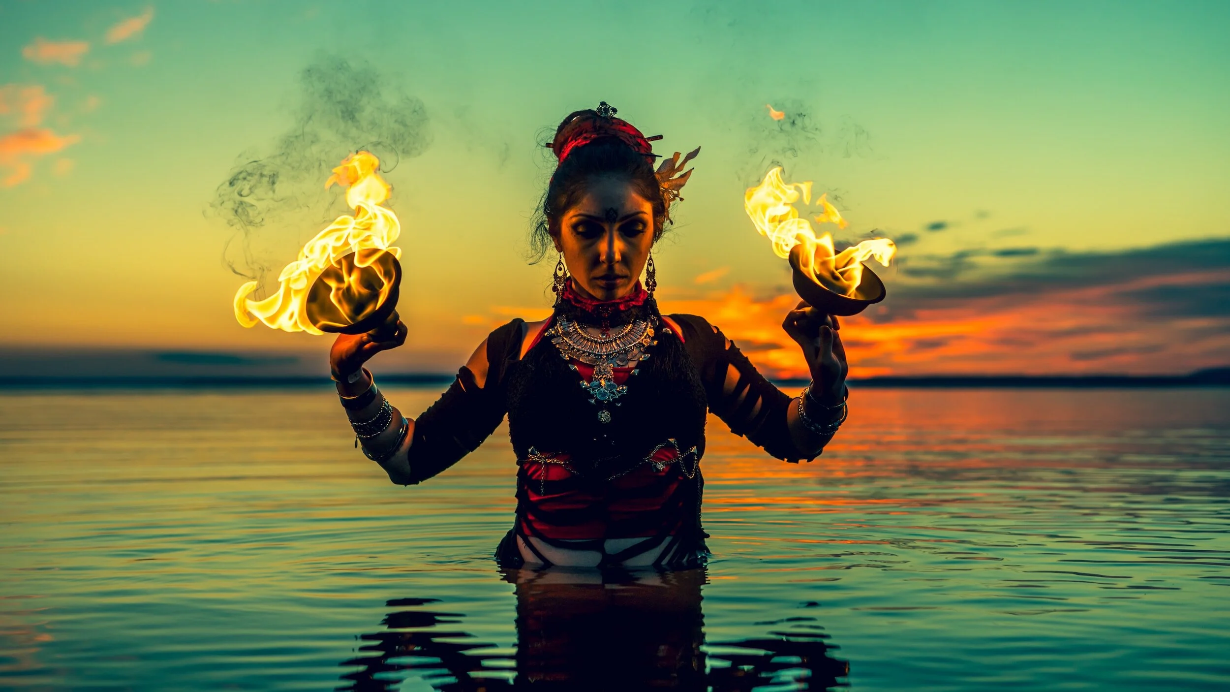 A woman in traditional attire holding two flaming bowls during sunset at a lake.