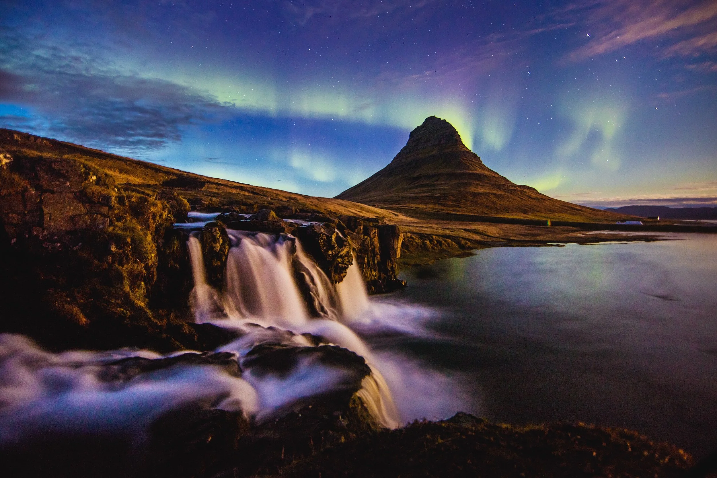 Nighttime scene of Kirkjufell mountain with the Northern Lights in the sky, a waterfall in the foreground, and a calm body of water reflecting the landscape.