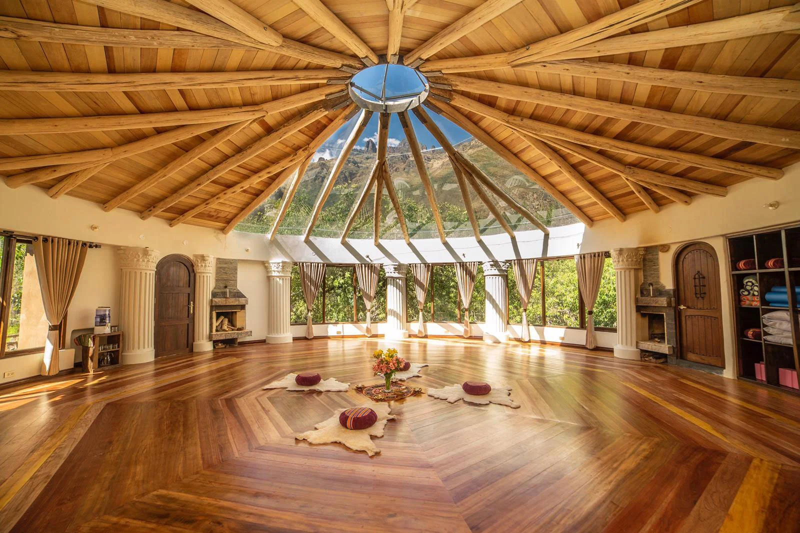 Spacious, round yoga or meditation studio with polished wood floors, high wooden ceiling with skylight, large windows with curtains, and cozy floor cushions with a flower arrangement in the center.