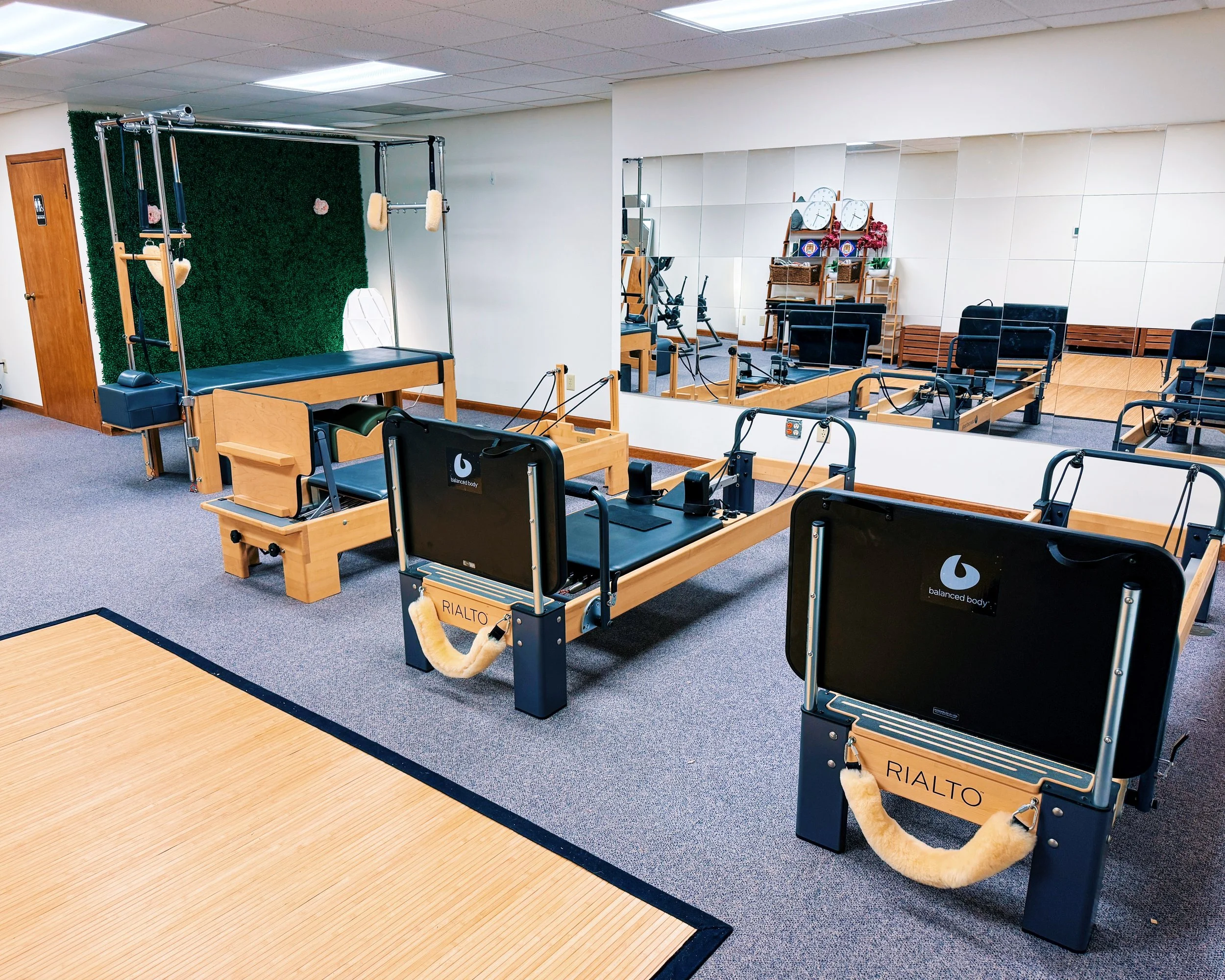 Physical therapy room with specialized equipment, including two RIALTO balanced body reformer machines, a vertical wall with mirrors, and a green textured wall in the corner.