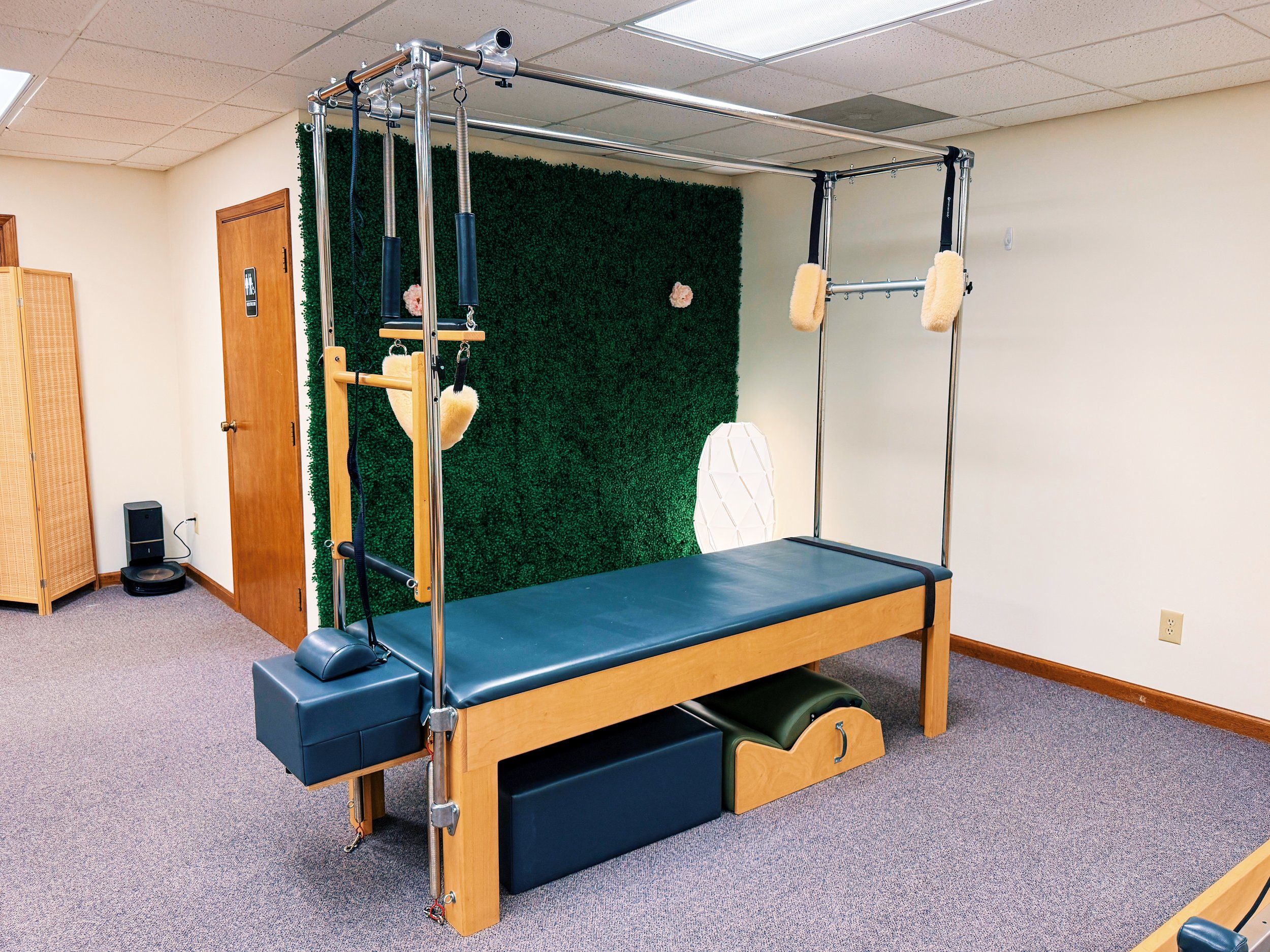 A Pilates reformer machine with various attachments in a room featuring a green artificial grass wall and beige walls.