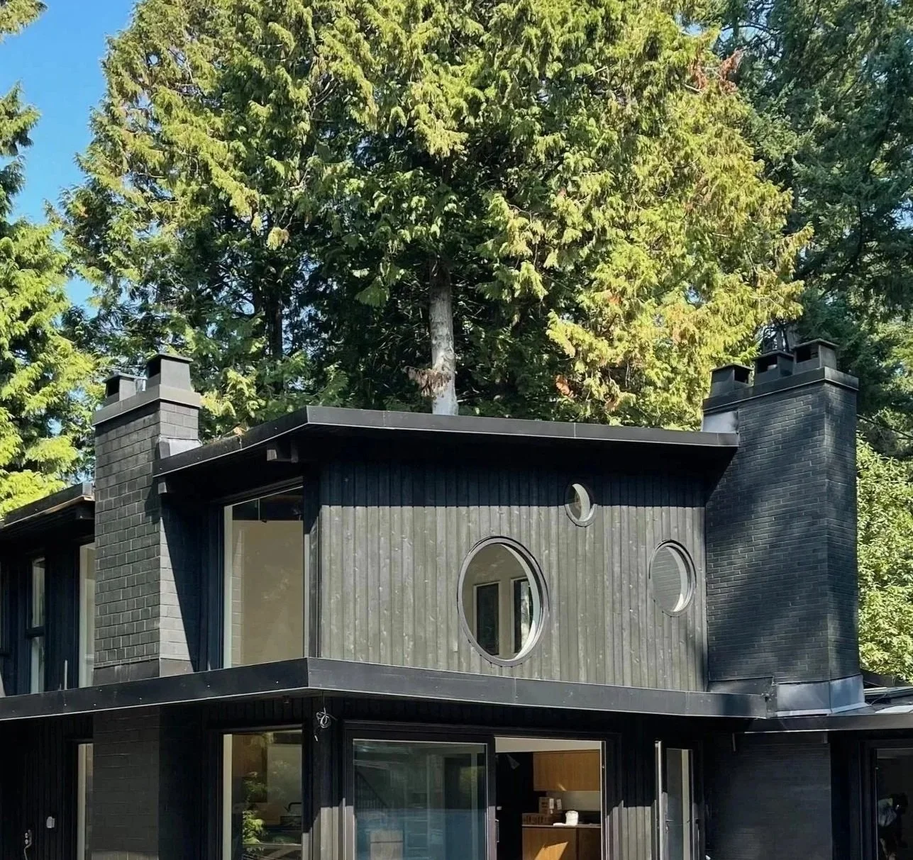 Contemporary renovation to a mid century home in Crescent Beach BC with black exterior, round windows, large glass sliding doors, surrounded by tall trees, under clear blue sky.