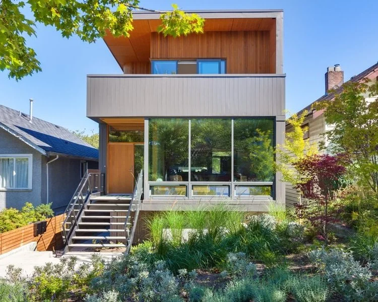 Modern two-story house with large glass windows, wooden accents, and a staircase leading to the entrance, surrounded by greenery and trees.