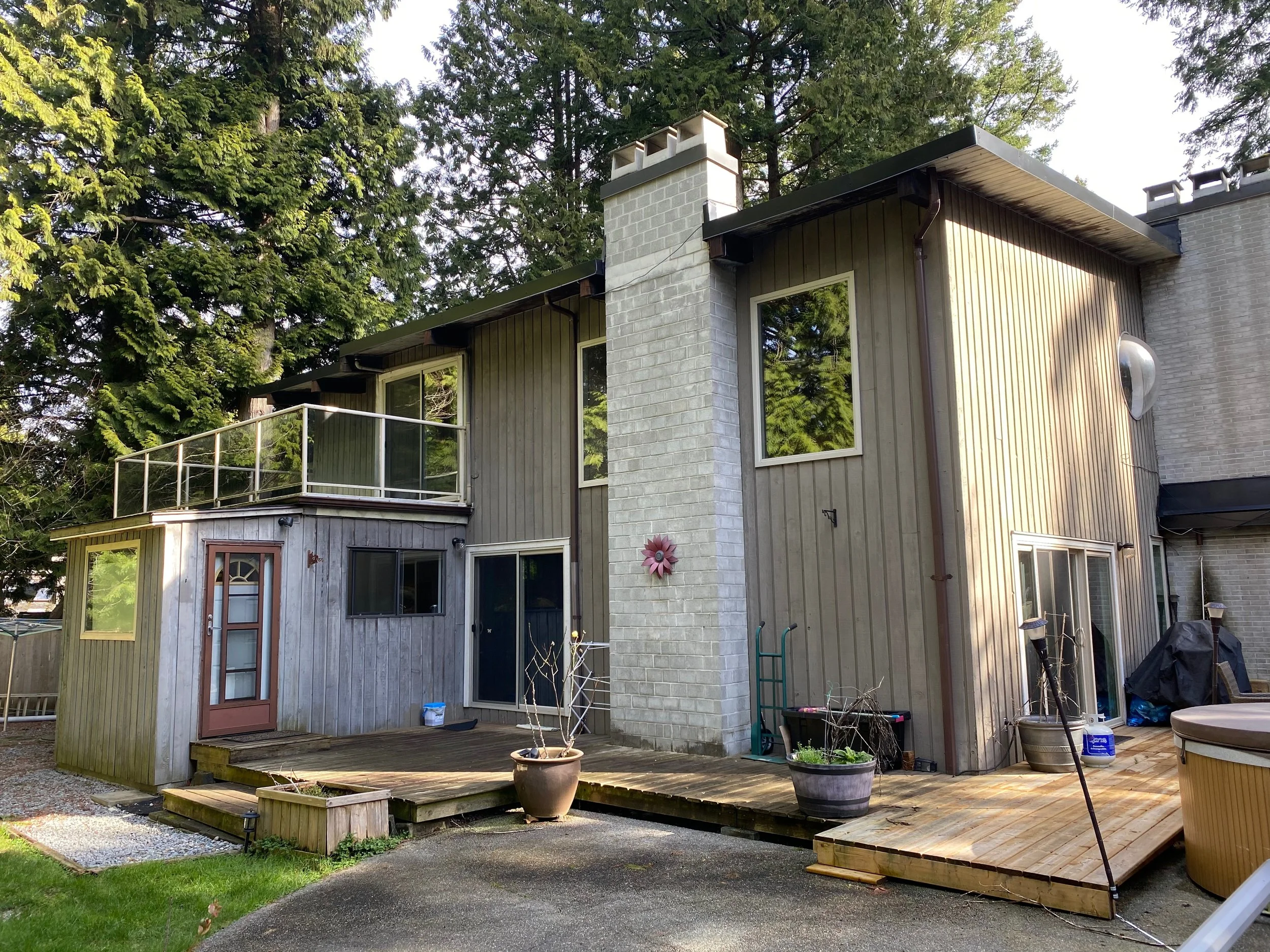 The back of a two-story house with a wooden deck, potted plants, and trees in the background.