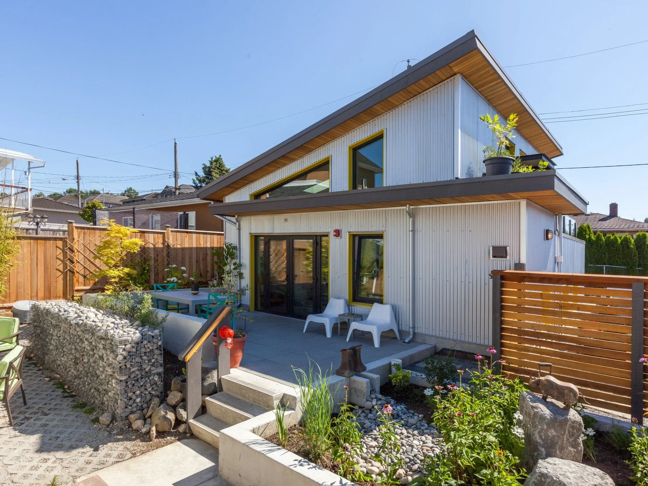 Modern two-story house with large windows, a small patio with chairs, a wooden fence, and a garden with plants and flowers.