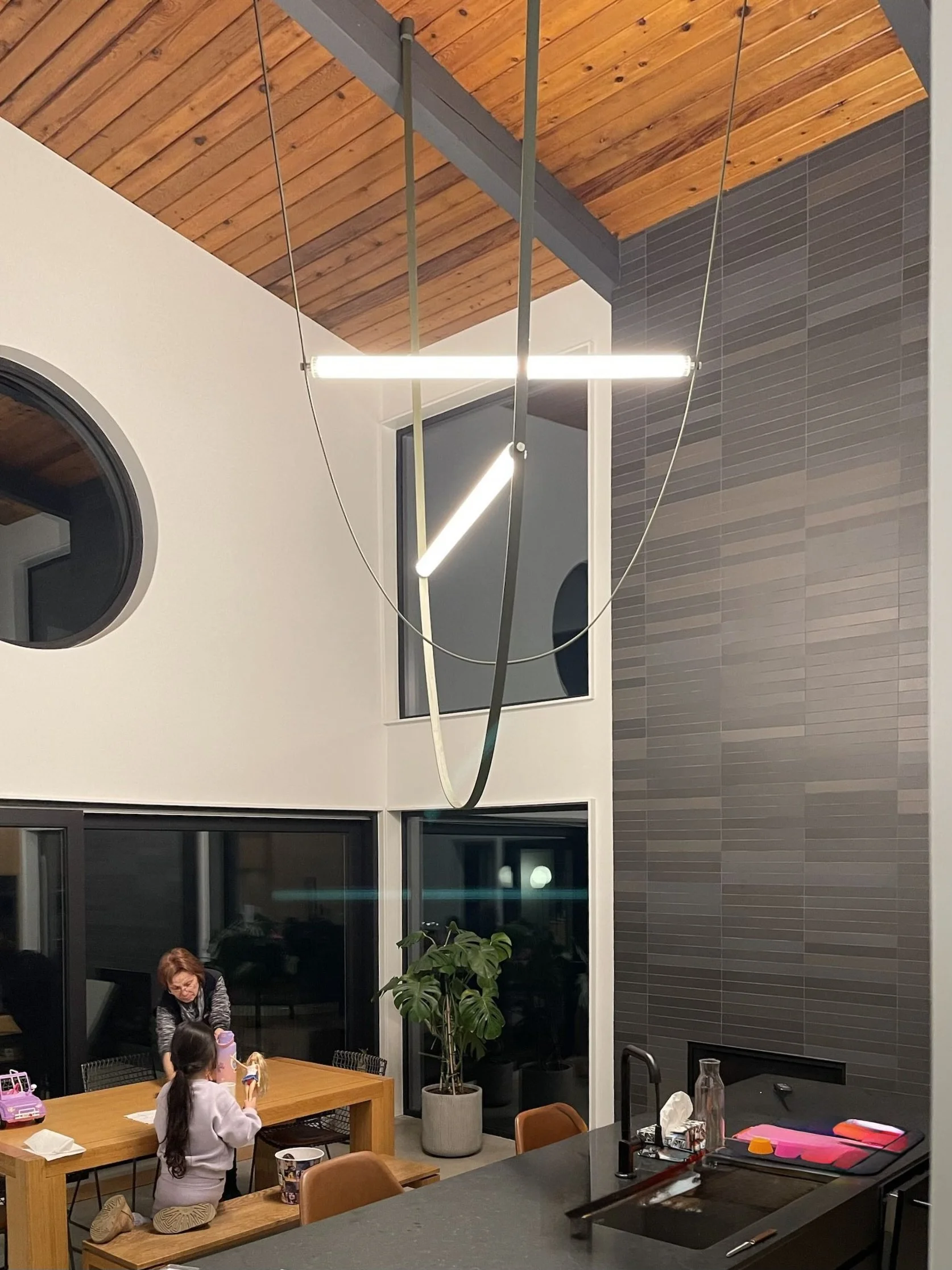 Interior of a modern home with high vaulted wood ceiling, black tiled accent wall, and large windows. Two women, one adult and one child, are at a wooden dining table with toys. A large green plant is beside the windows, and kitchen items are on the counter.