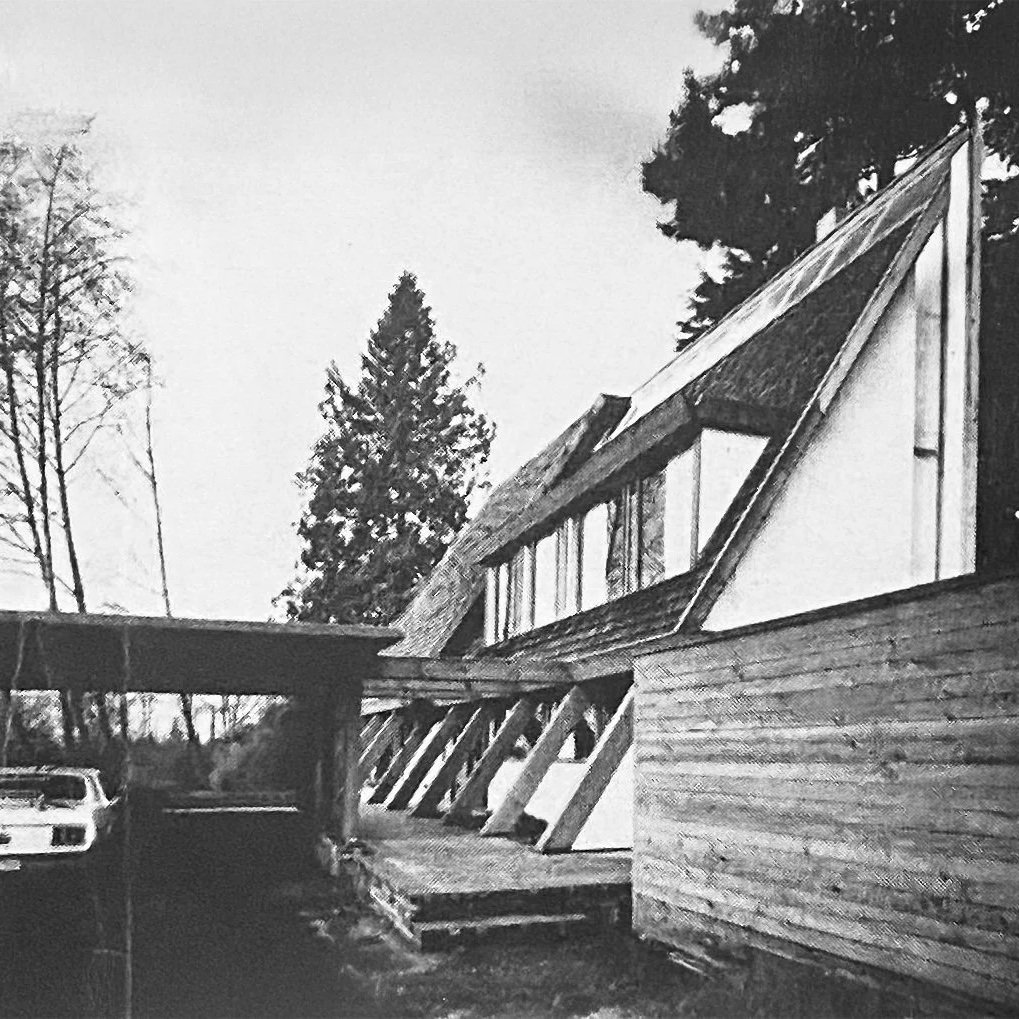 Black and white photo of the Wedge house by Arthur Erickson and a wooden exterior wall, surrounded by tall trees. An overturned vehicle is parked underneath a carport.