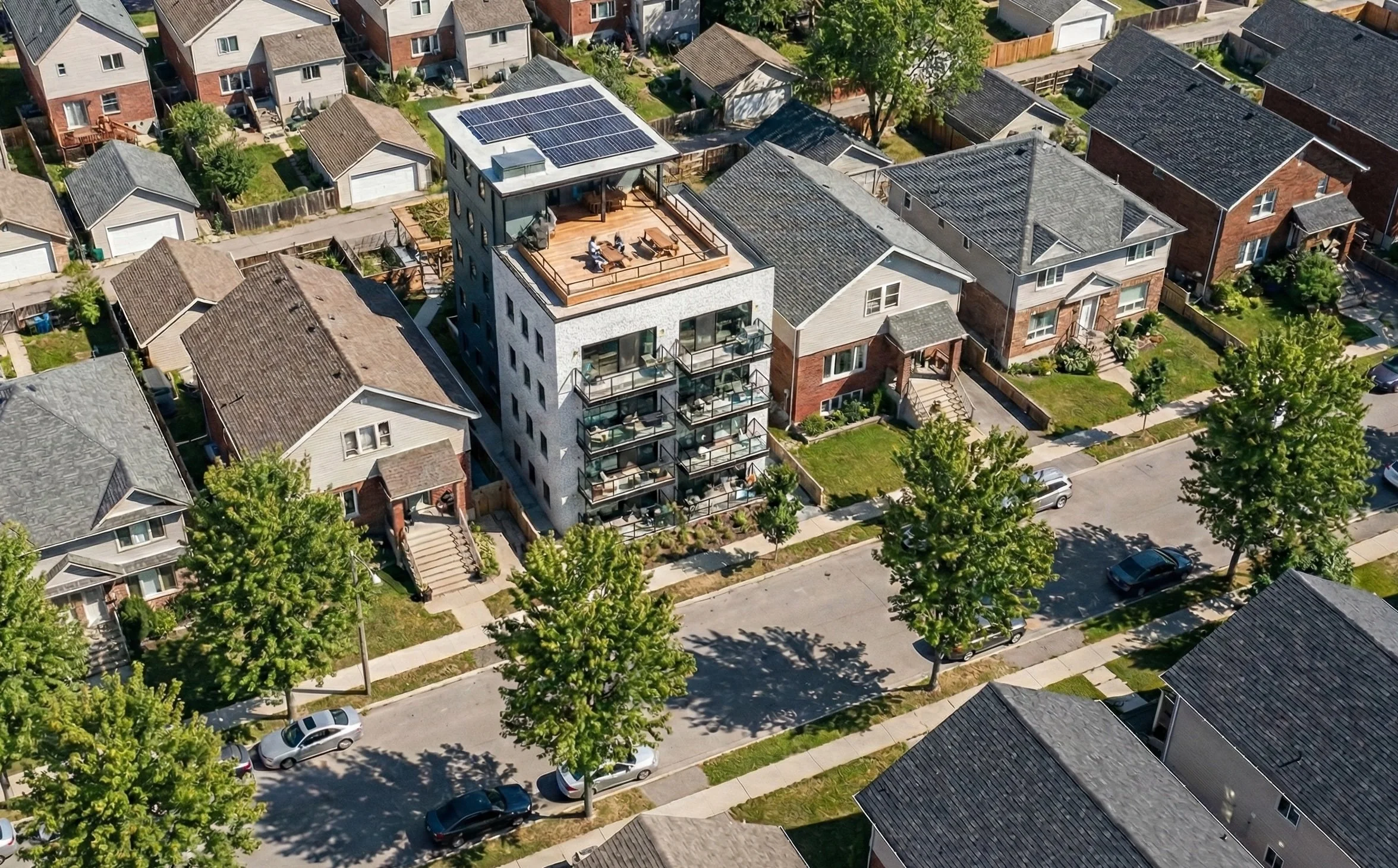 Aerial view of a residential neighborhood showing a modern multi-story apartment building with a rooftop terrace, solar panels, and several balconies, surrounded by traditional houses with trees and parked cars on the street.