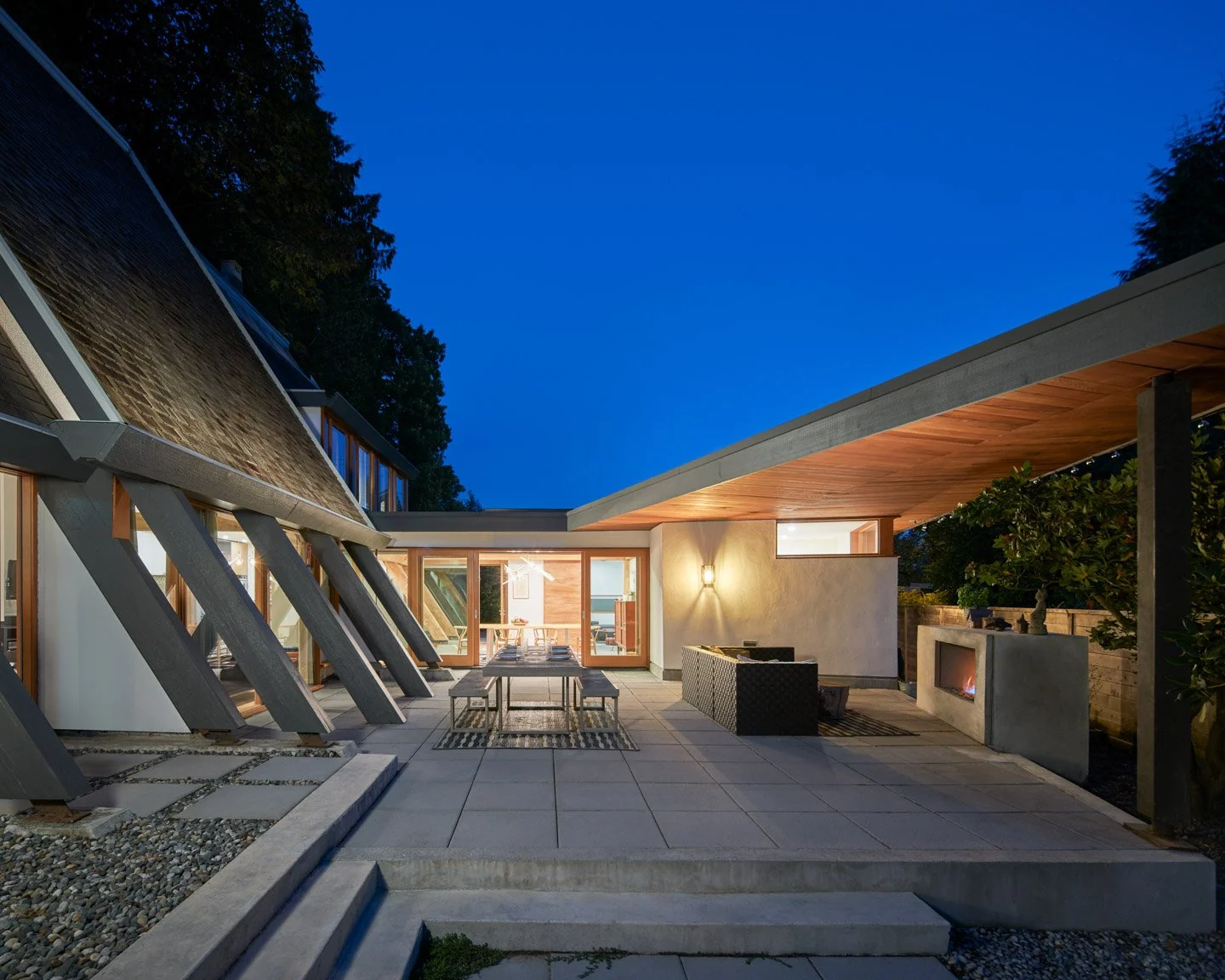Lanefab's renovation of the Wedge House by Arthur Erickson, patio at dusk with seating, fireplace, and outdoor lighting under a wooden roof extension.
