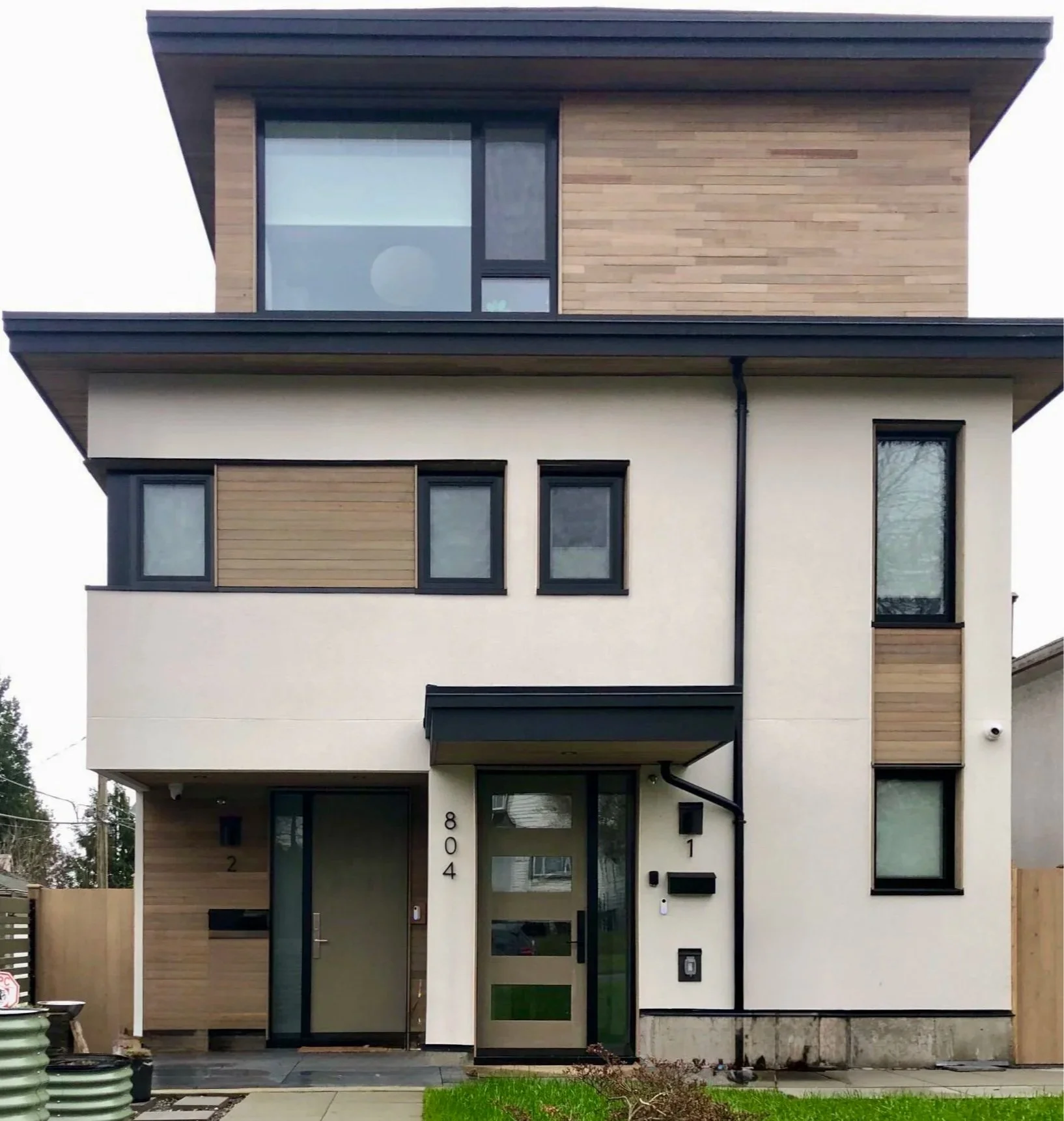 A modern multi-story house with a flat roof, featuring a combination of white walls, black window frames, and wooden panels. The house has multiple windows of different sizes and a front door with glass panels. There are small patches of green grass and a paved walkway in front.