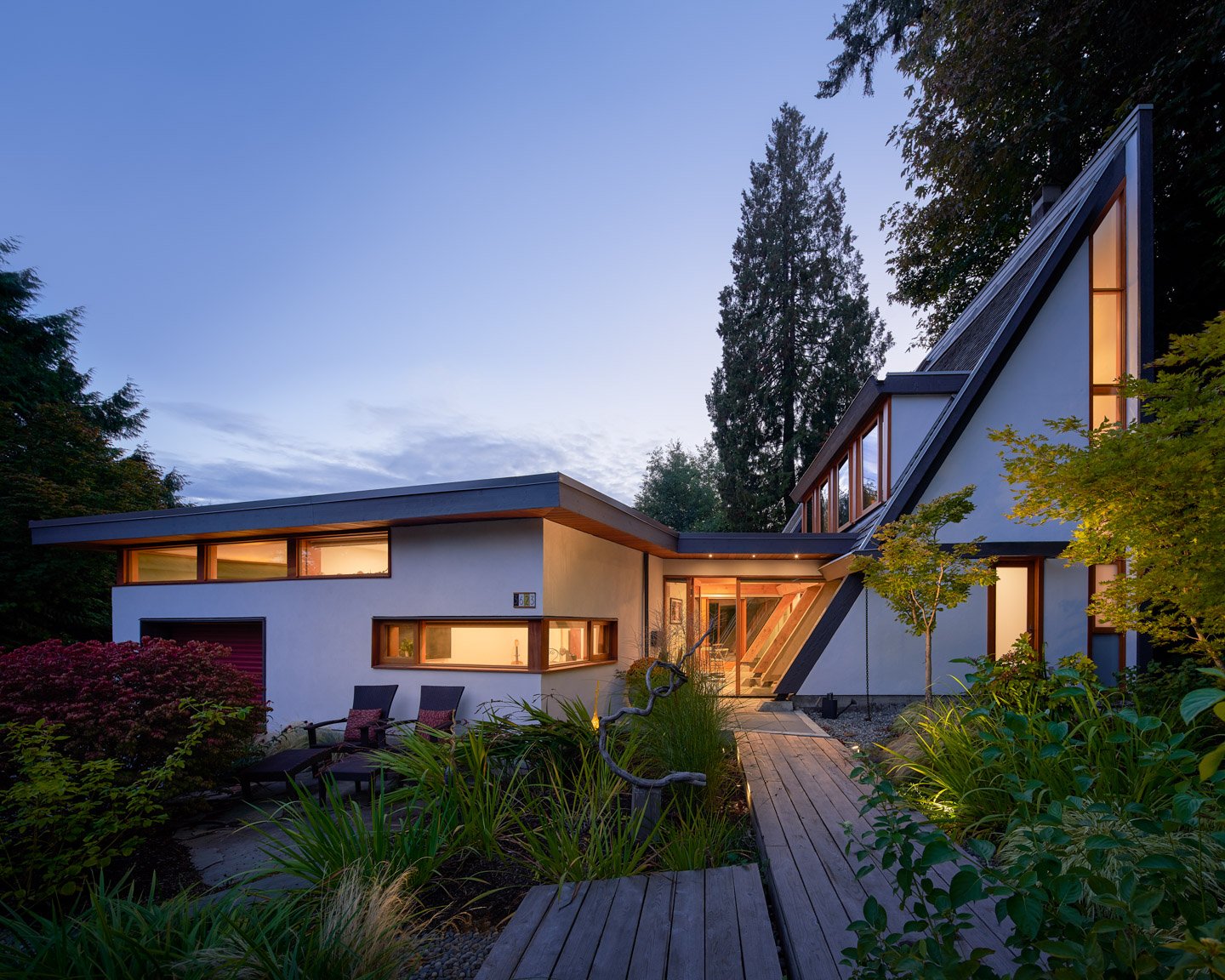 Lanefab's renovation of the Wedge House by Arthur Erickson with large windows surrounded by greenery at dusk, with warm interior lighting visible through the windows.