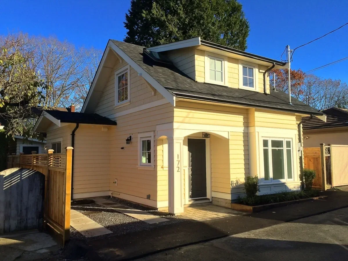 A two-story yellow house with white trim, a small covered front porch, and black front door, located in a sunny neighborhood with blue sky and trees in the background.