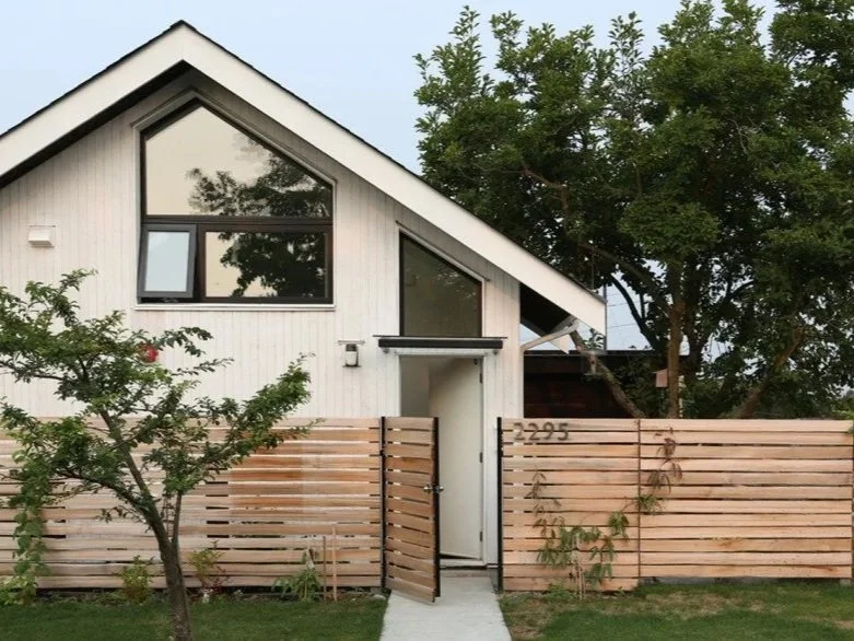 Modern two-story house with a sloped roof, large windows, and a wooden fence in front. There are trees and a sidewalk leading to the front door.