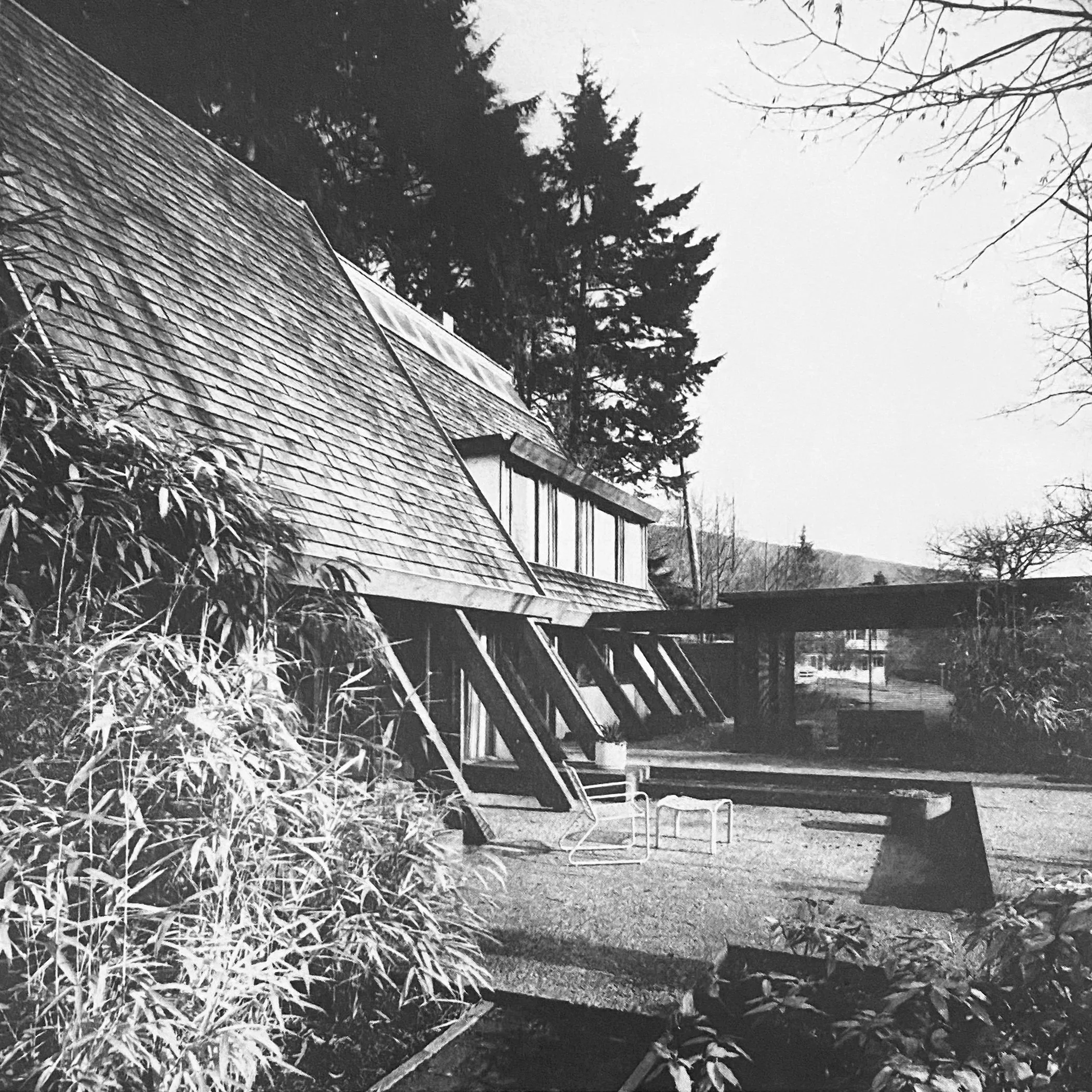 Black and white photo of the Wedge House by Arthur Erickson with large windows and sloped roof, surrounded by trees and outdoor furniture, on a clear day.