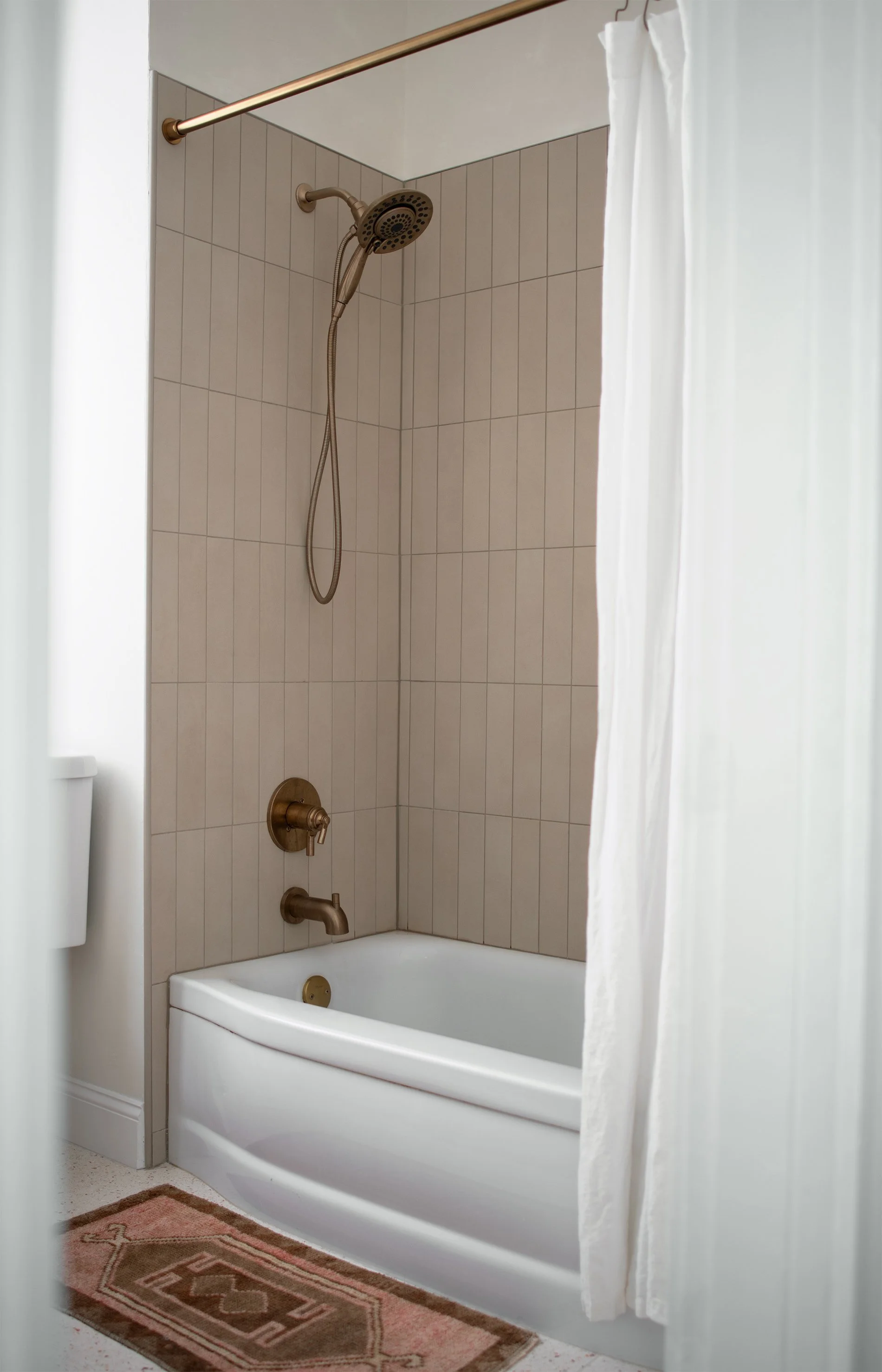 Tan vertical shower tile in rows with brass fixtures and a vintage pink turkish rug