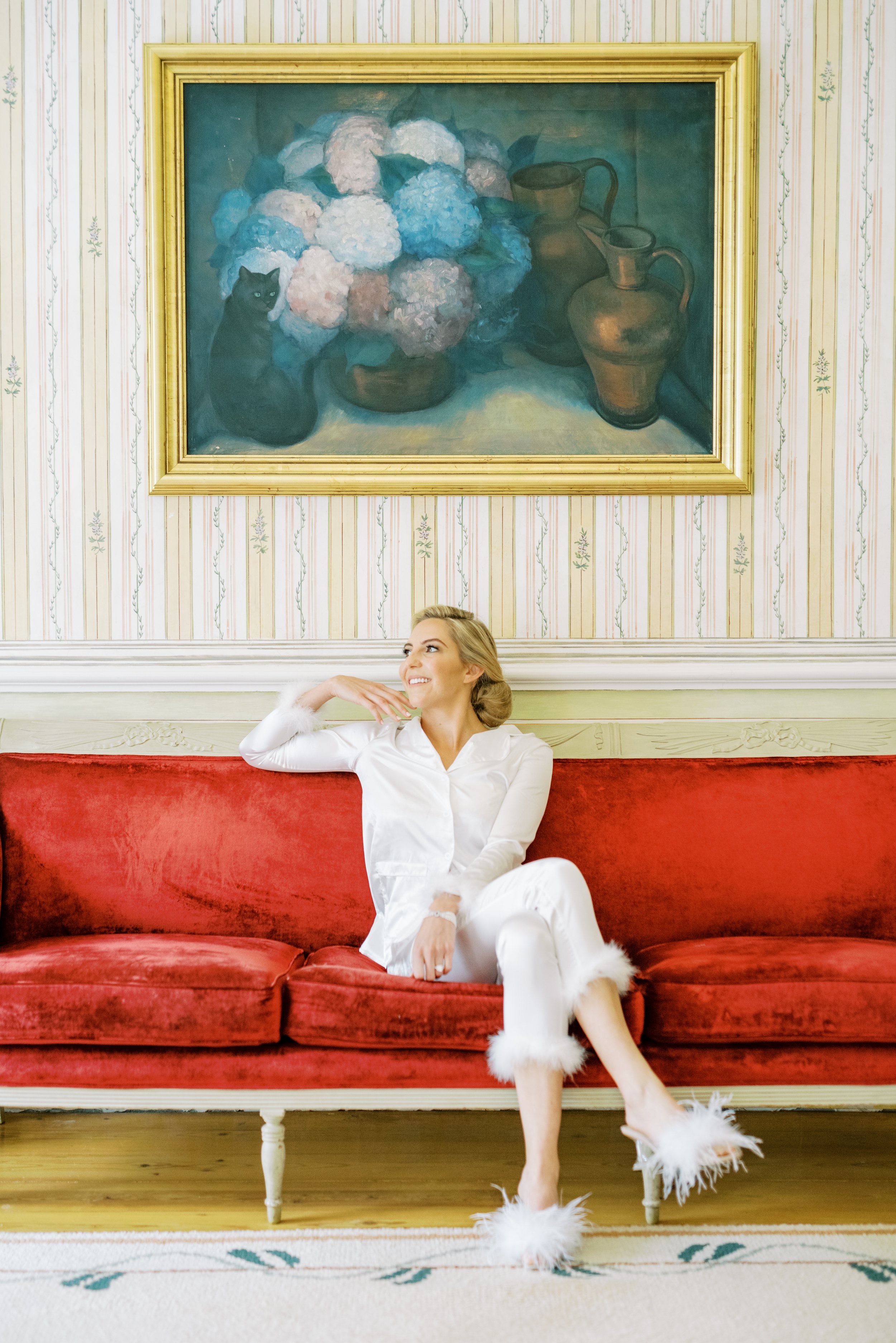 Bride in her white pijamas sitting on a luxurious red velvet sofa and an antique painting behind her