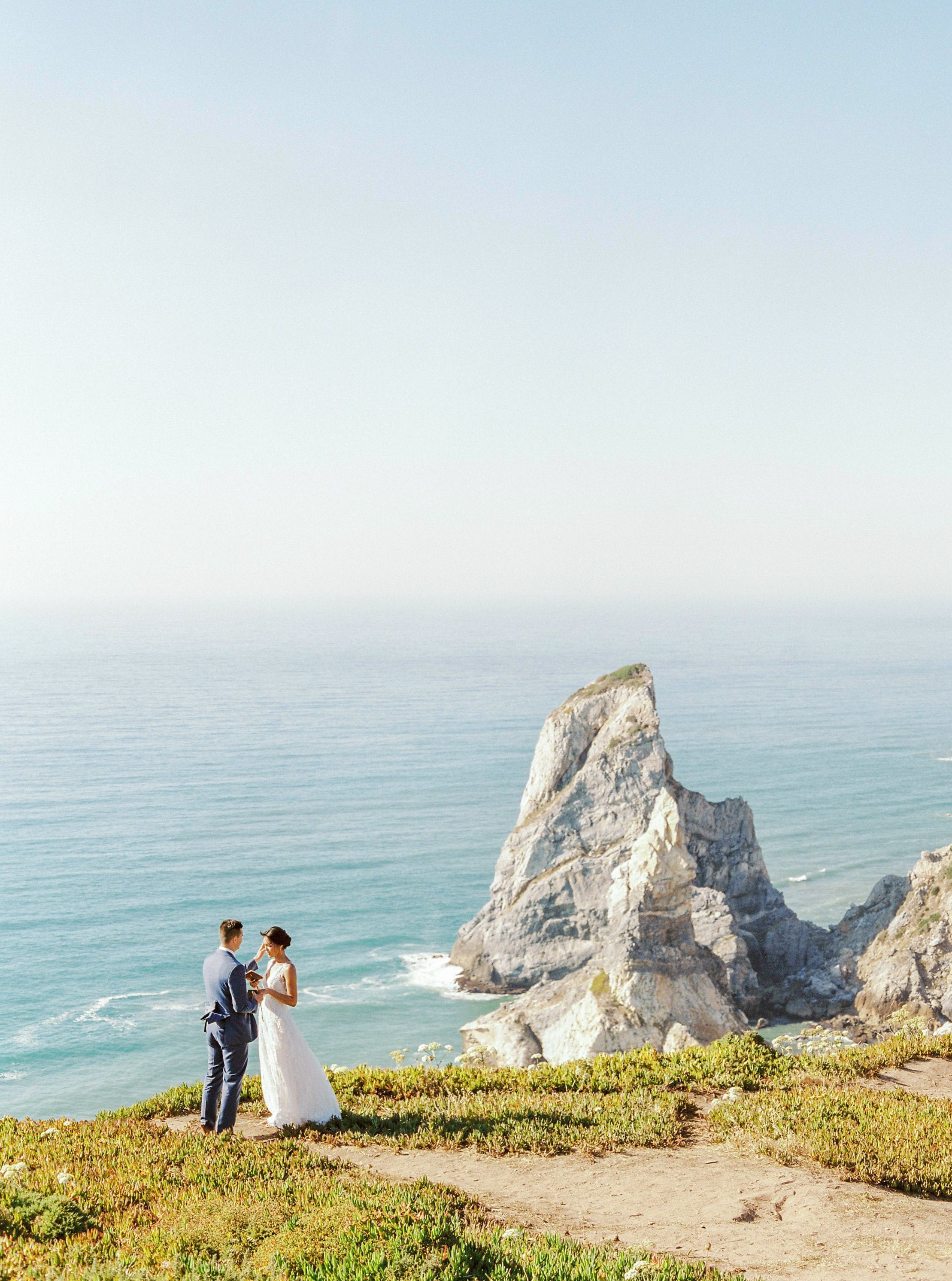 Bride and Groom standing on top off the cliff at Praia da Ursa with the blue ocean and sunny sky as a backdrop