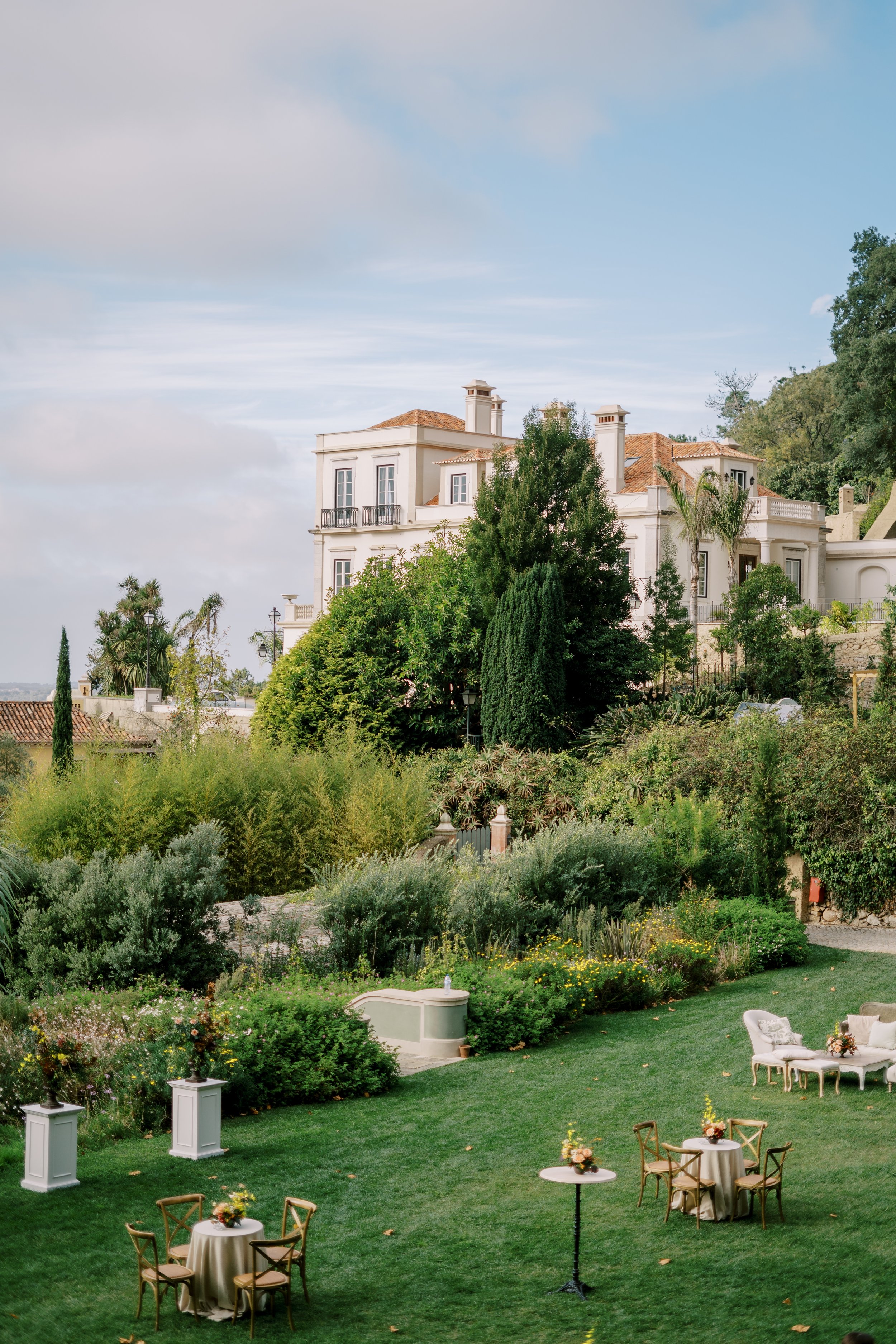 A photo of Quinta da Bela Vista in Sintra, captured from the garden, the building is partially hidden by tall trees, and the tables are already set for the cocktail hour in the garden