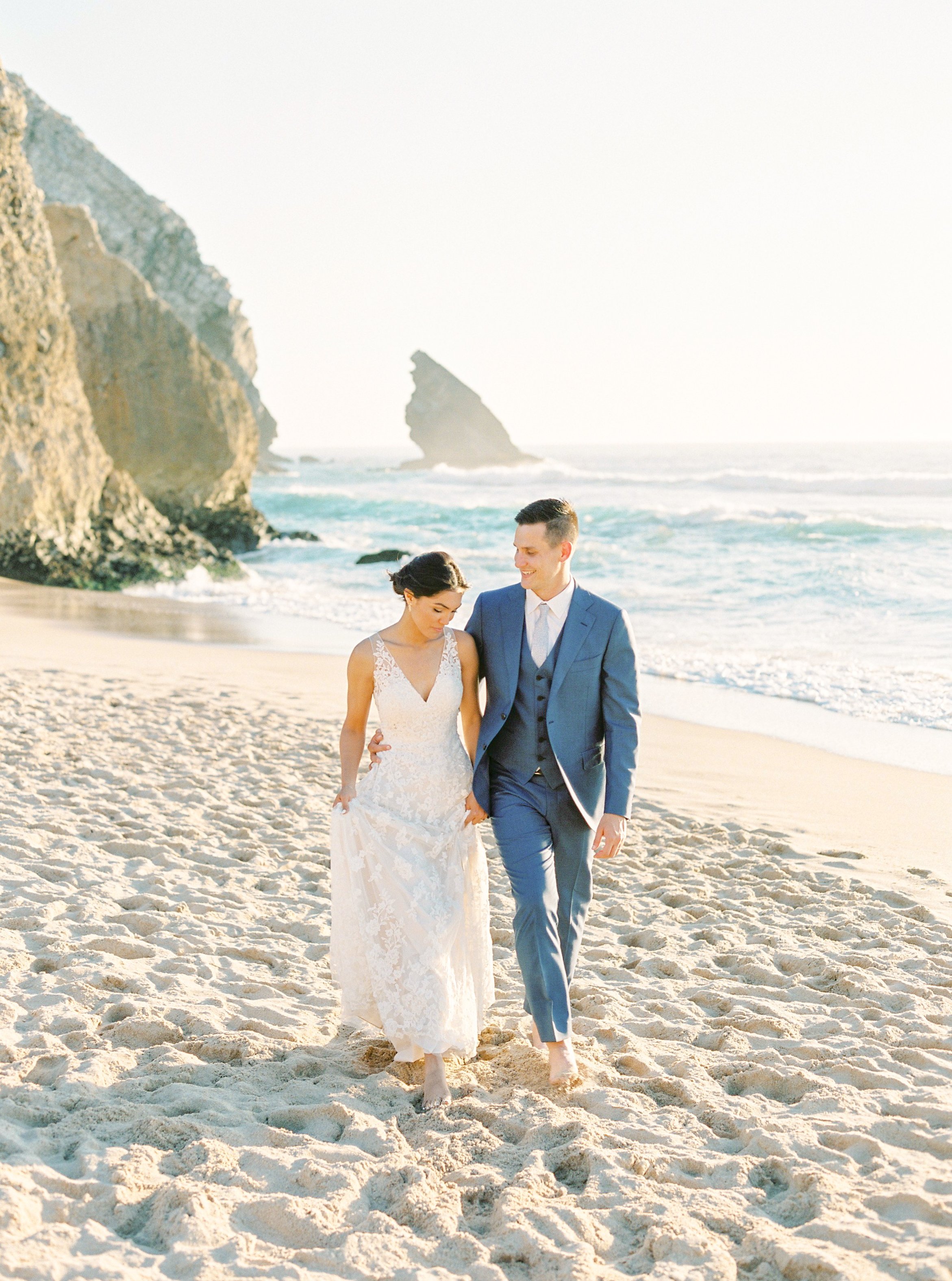 Bride and groom walking hand-in-hand on the beach of Praia da Ursa with impressive cliffs and wavy ocean in the background