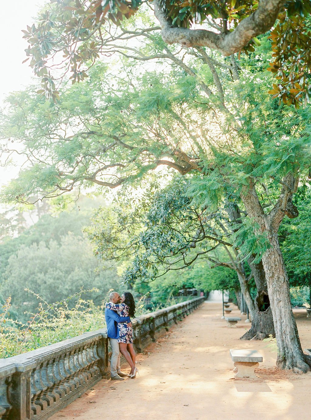 An engagement photo of a couple hugging on the pathway in a park, with tall trees surrounding them