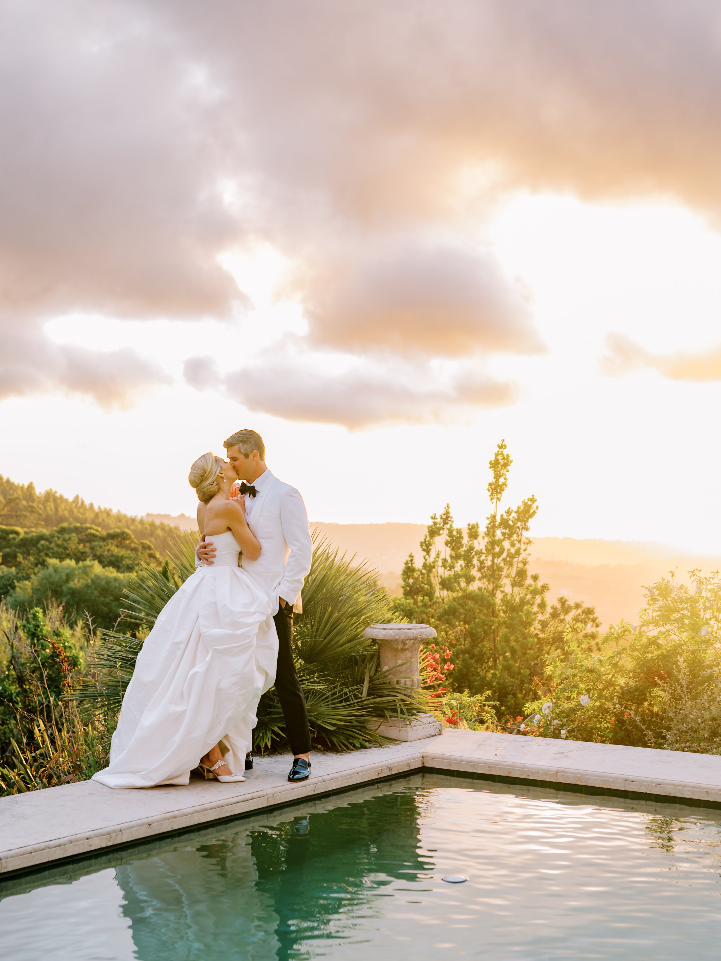 Bride and groom kissing at the edge of the pool at Quinta da Bela Vista with the view of the Sintra hills and the setting sun in the background, painting everything in an orange-yellow-purple color