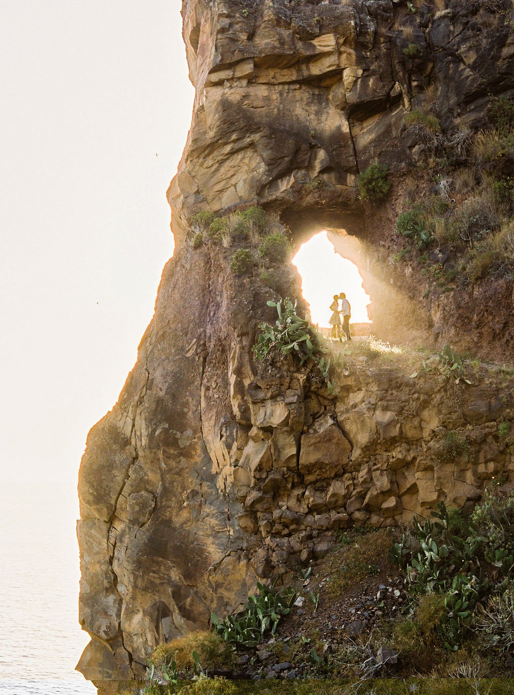 A breathtaking image of an arche formed withing a cliff on the side of the mountain in Madeira, with a couple kissing underneath while the light of the setting sun is coming through