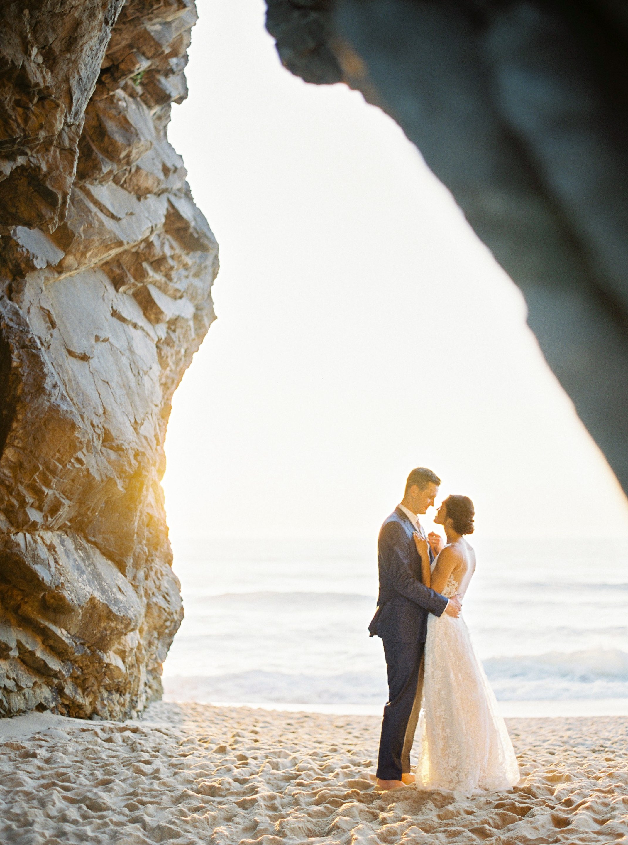 Bride and groom standing under the arche formed by a cliff right next to the ocean, while the light of the setting sun comes through