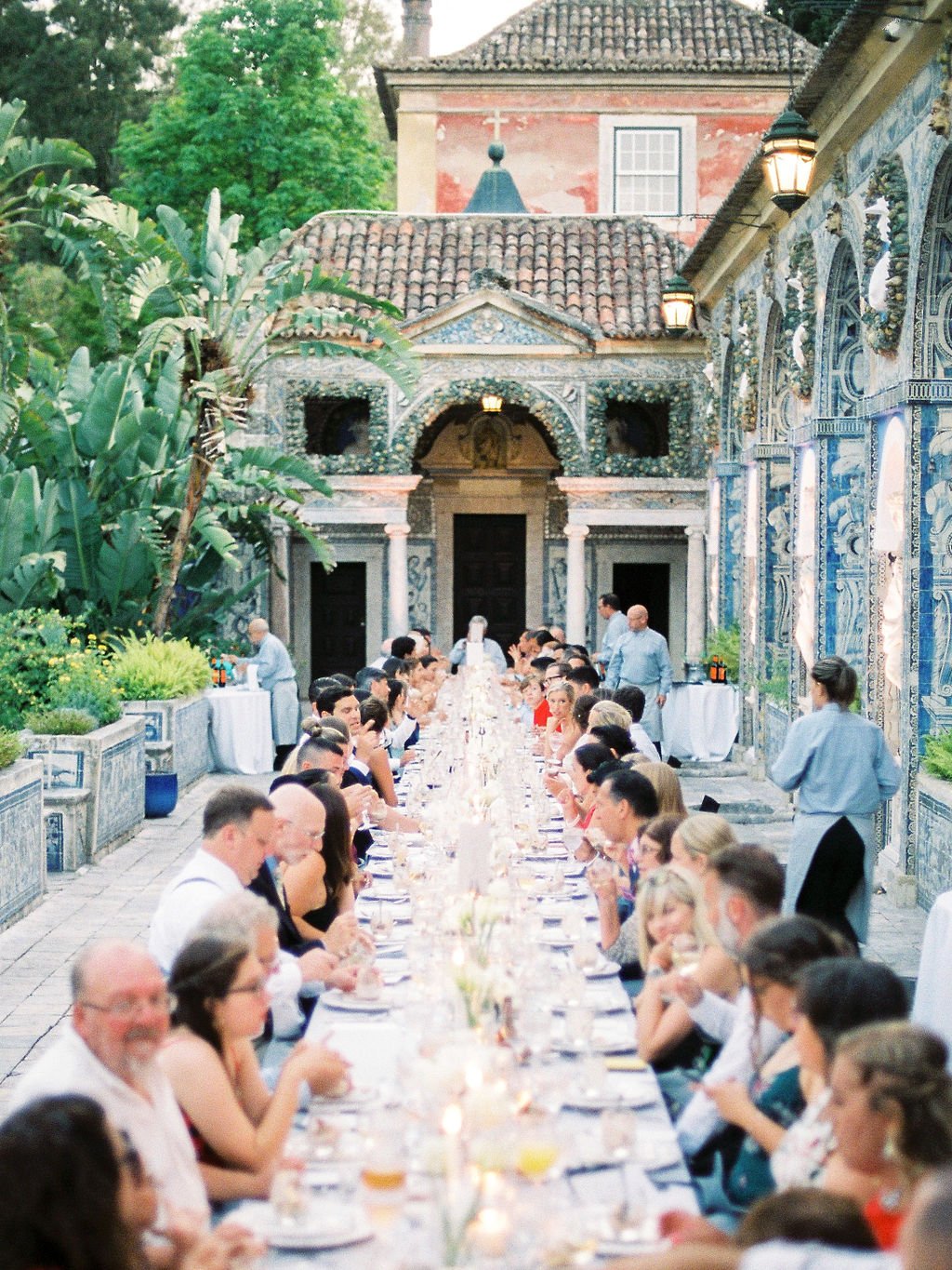 A long, beautifully set table in the terrace of  the Marqueses de Fronteira Palace in Lisbon with Portuguese tiles as backdrop, while the guests are chatting and eating