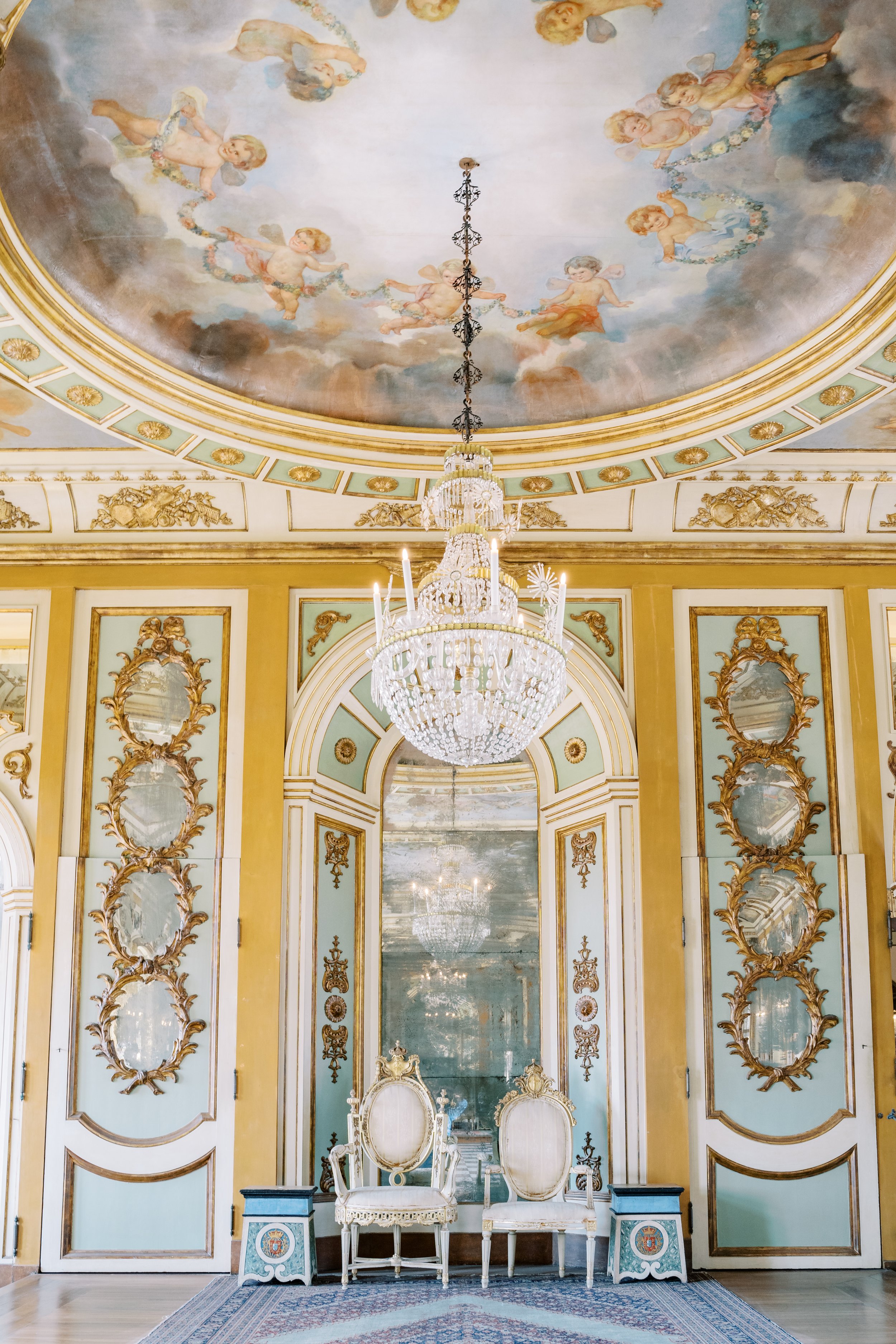 The interior of Palácio de Queluz showing an impressive chandelier, a mirror in the middle, with two chairs on the both sides and a beautiful painting on the ceiling