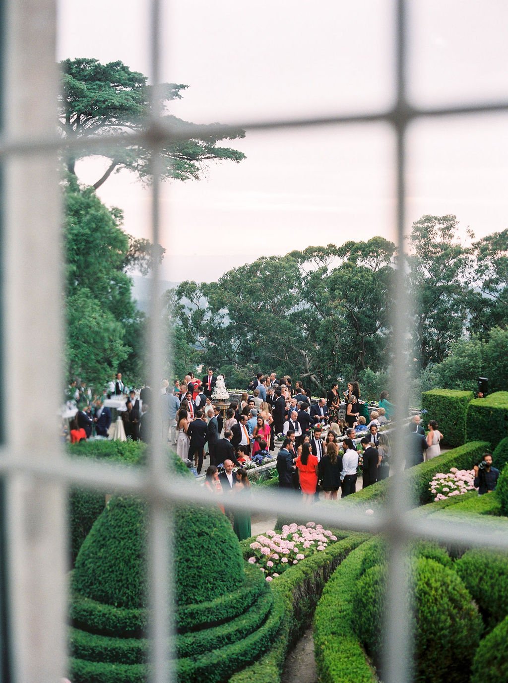 Photo from inside Palácio de Seteais though a window, showing wedding guests gathered under the tall trees in the garden for cocktail hour