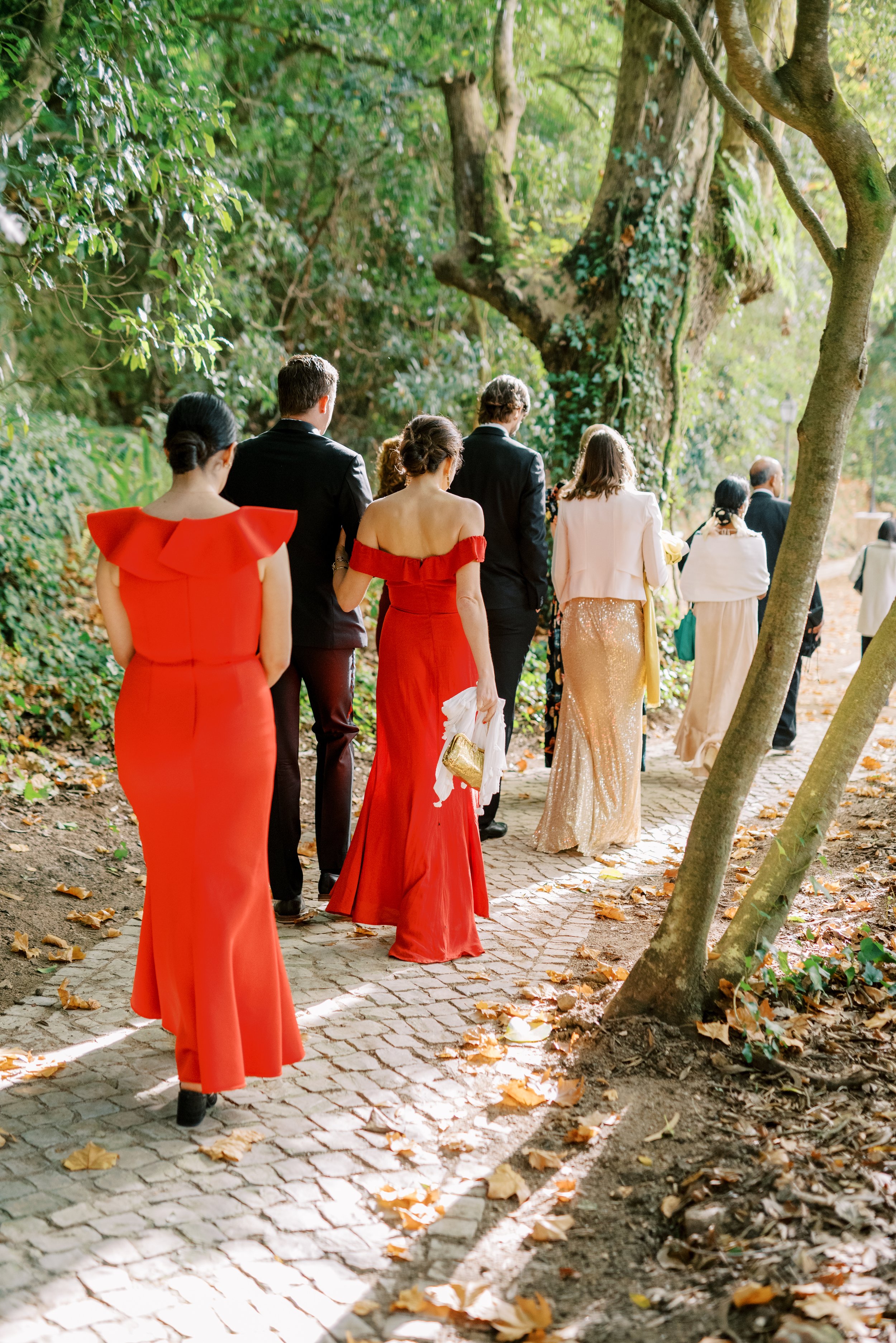 The wedding delegation walking on the path towards the ceremony area between the trees, dressed in elegant, red dresses