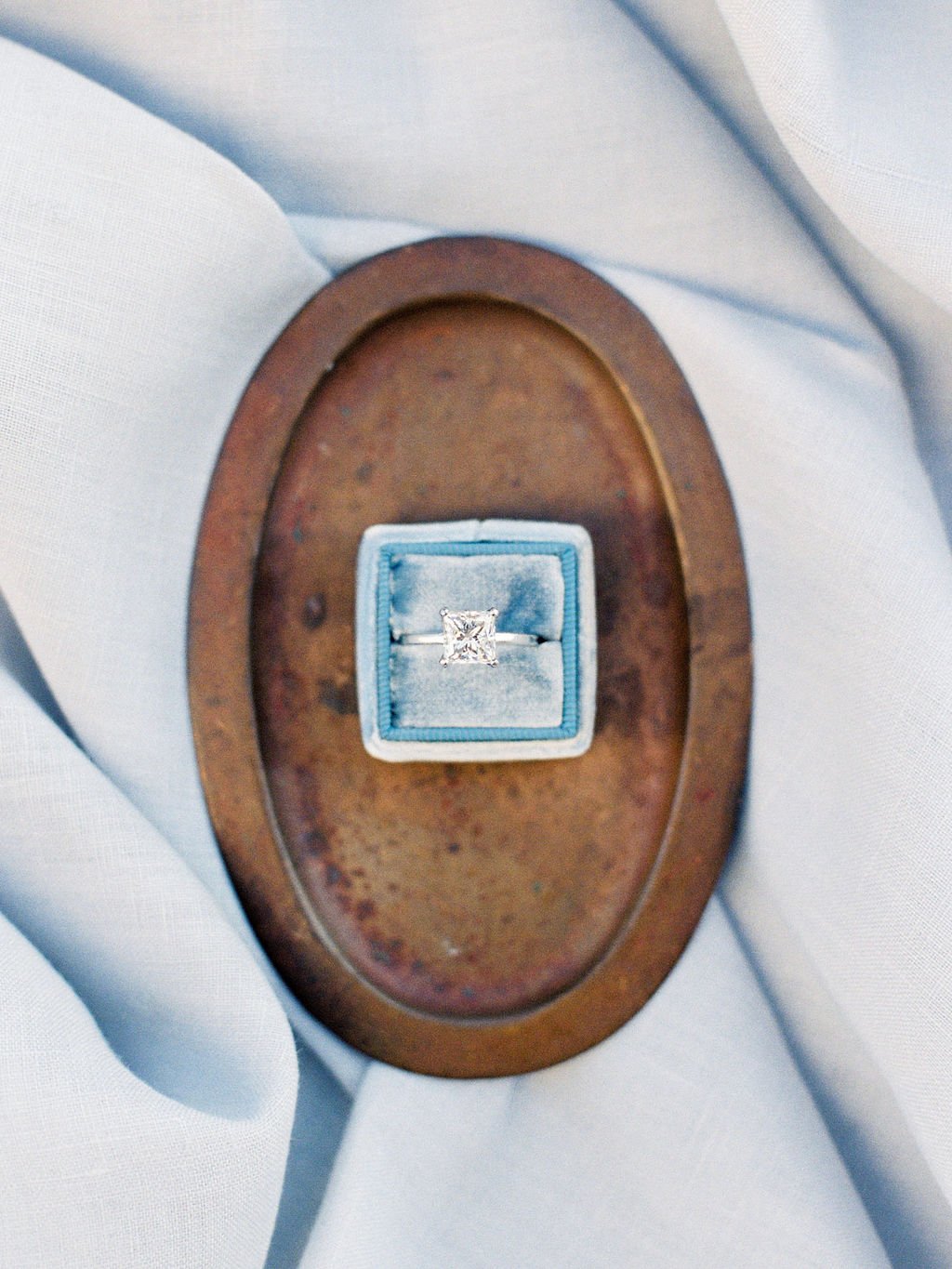 A square diamond wedding ring in it's blue box captured from above on a brown oval plate