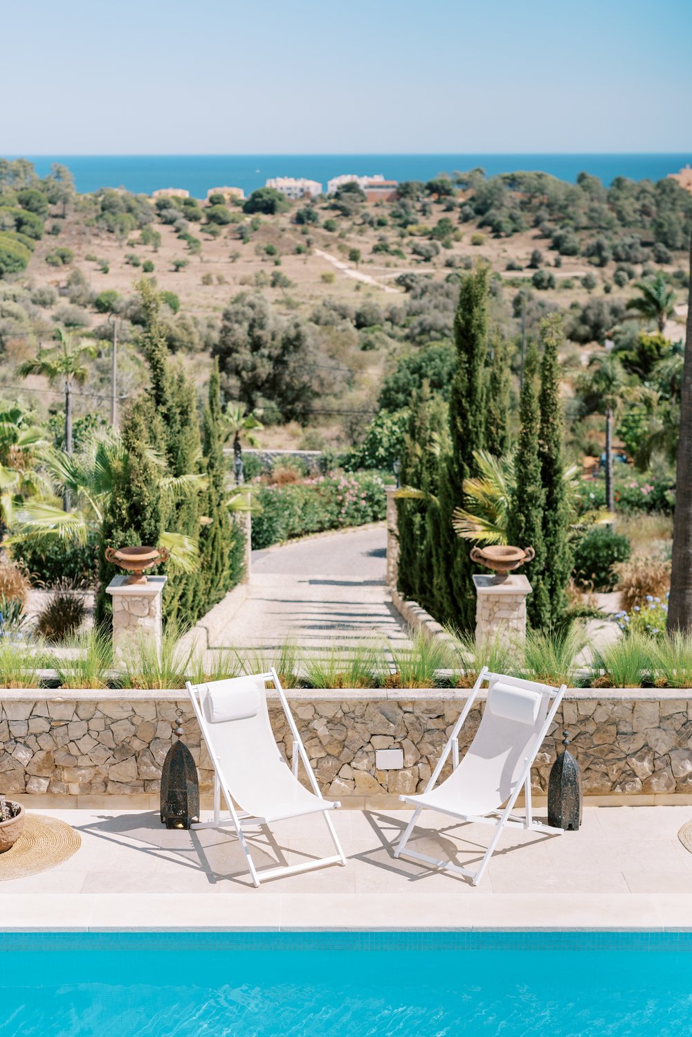 Photo showing the edge of a swimming pool, two chairs above and the blue ocen and sky as a backdrop, a typical view from the Algarve, South of Portugal, in a hot, summer day