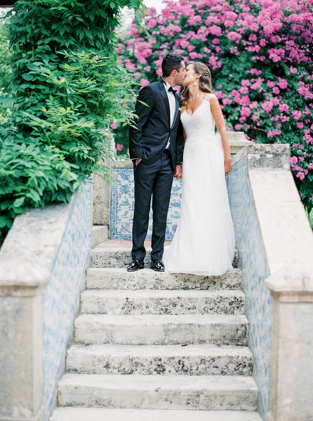 Bride and groom standing on top of the stairs outside a Portuguese building decorated with blue and white tiles and with pink flowers in the background