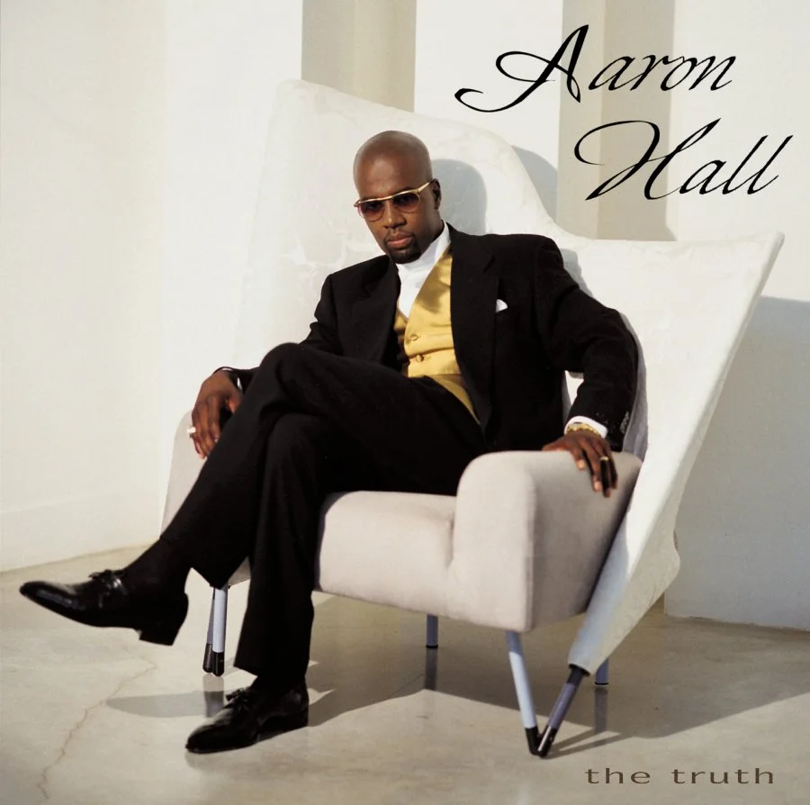 Aaron Hall sitting in a white chair while wearing a suit and looking hot.jpg