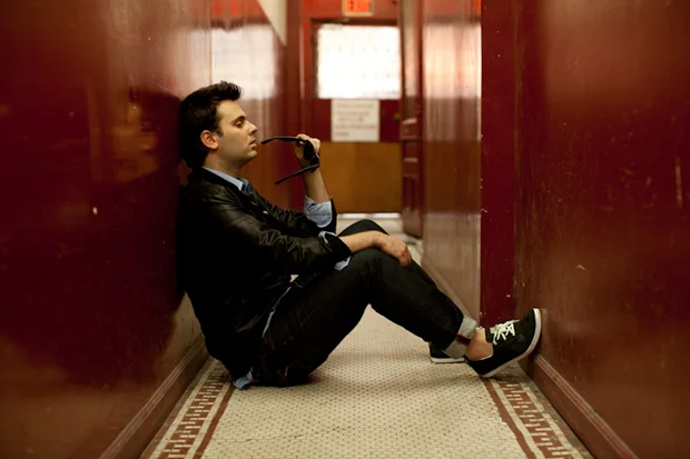 Outasight sitting on the floor in an apartment building's hallway with his sunglasses to his mouth.jpg