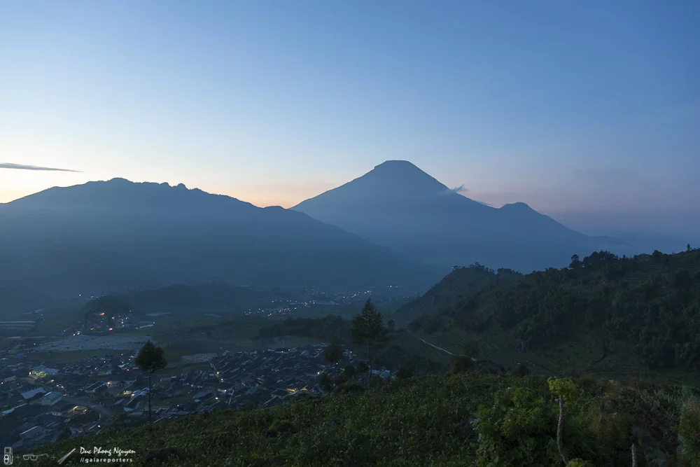 Sunset over a mountain range, with a prominent volcano in the background and a small town in the foreground.