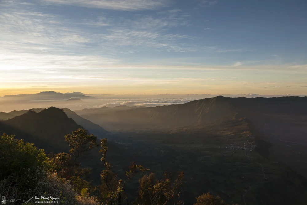Sunrise over a mountain range with lush green slopes, some trees in the foreground, and clouds hovering near the horizon.