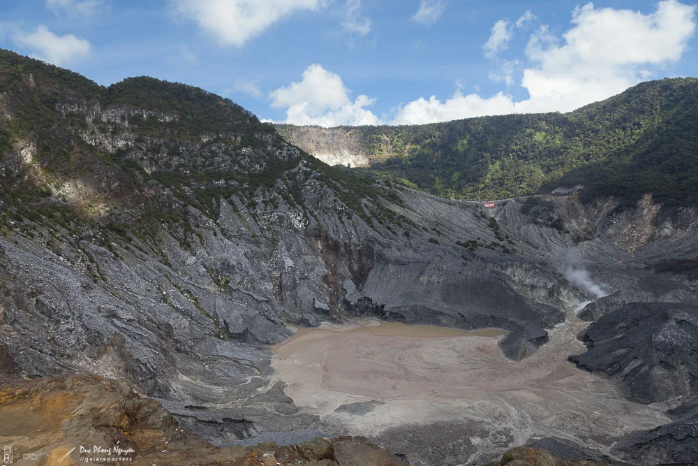 Volcano crater with dry, ash-covered ground and steep volcanic slopes, surrounded by green hills and partly cloudy blue sky.