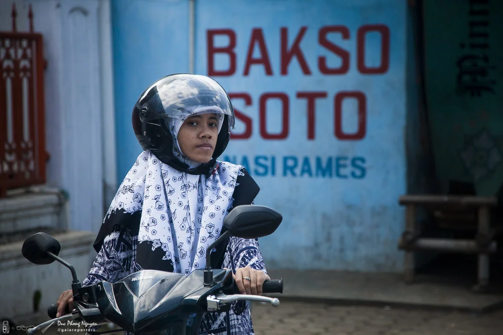 A woman wearing a helmet and scarf sitting on a black motorcycle in front of a blue wall with red and blue text advertising a restaurant or business.