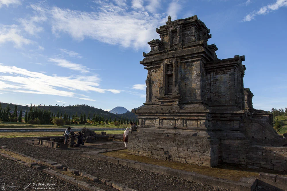 An ancient stone temple with a rectangular structure, set in a landscape with trees and mountains in the background, and a few people visiting.