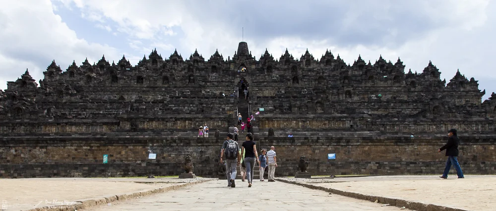 Group of tourists walking towards an ancient, large stone temple with multiple tiers and intricate architecture, under cloudy sky.